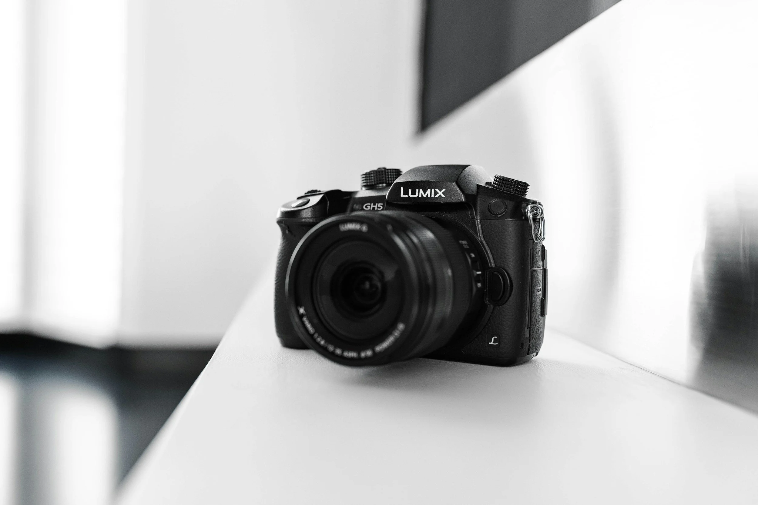 A black Panasonic Lumix GH5 camera with a lens attached, placed on a white surface with a minimalistic background.