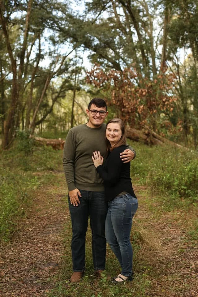 Owners standing close together on a wooded trail, embracing in a hug, surrounded by trees with green and brown leaves during daytime.