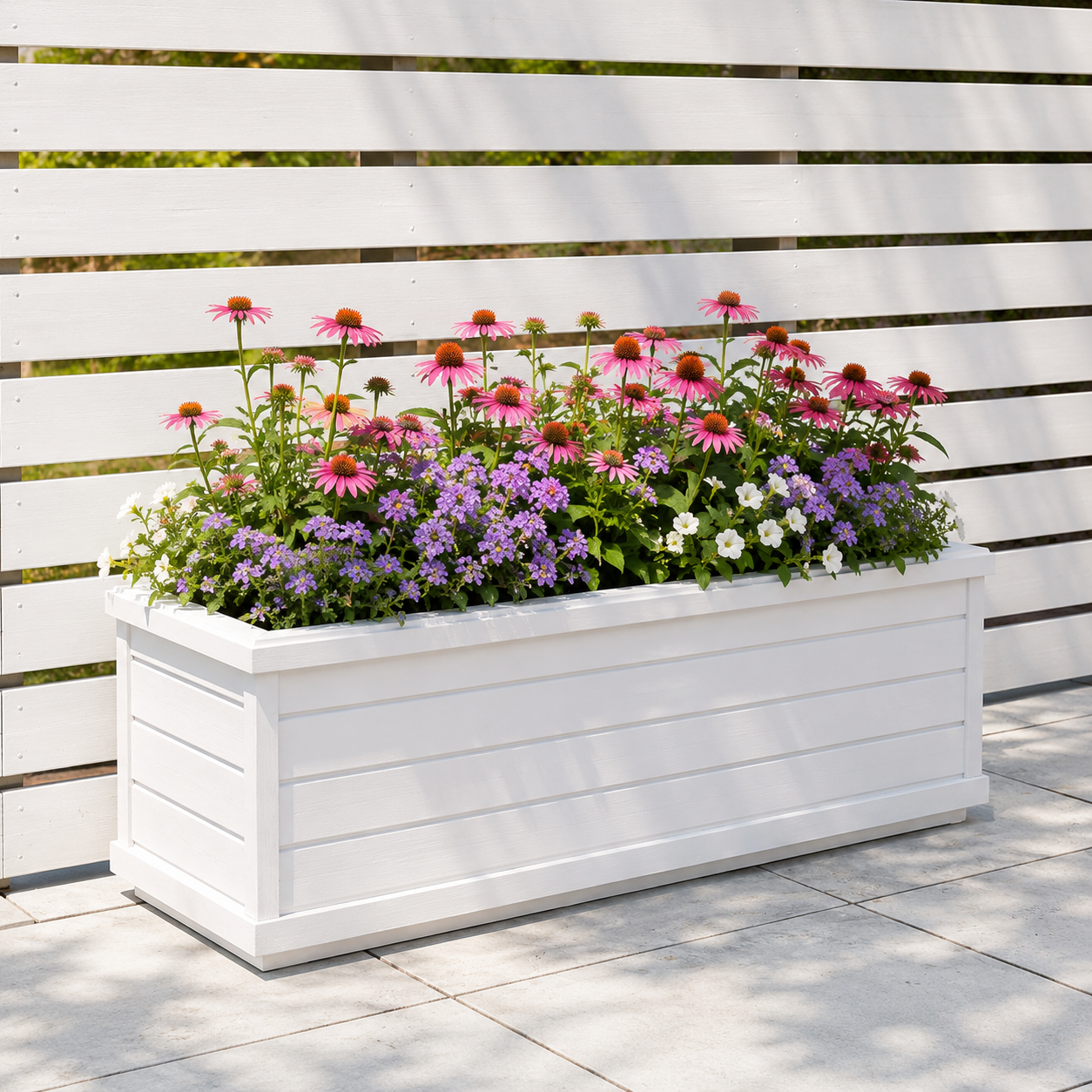 A large white flower planter with pink, purple, and white flowers in front of a white wooden slat fence.