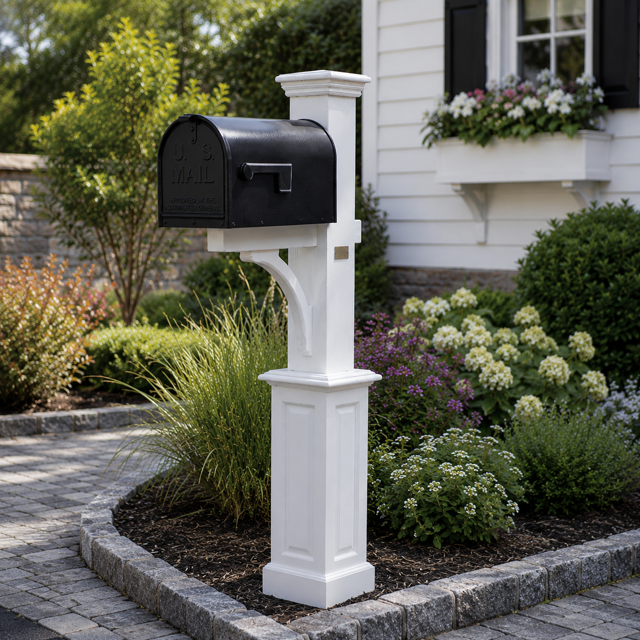 Black U.S. Mailbox mounted on a white post in a garden with various green shrubs and colorful flowers in front of a white house with black shutters.