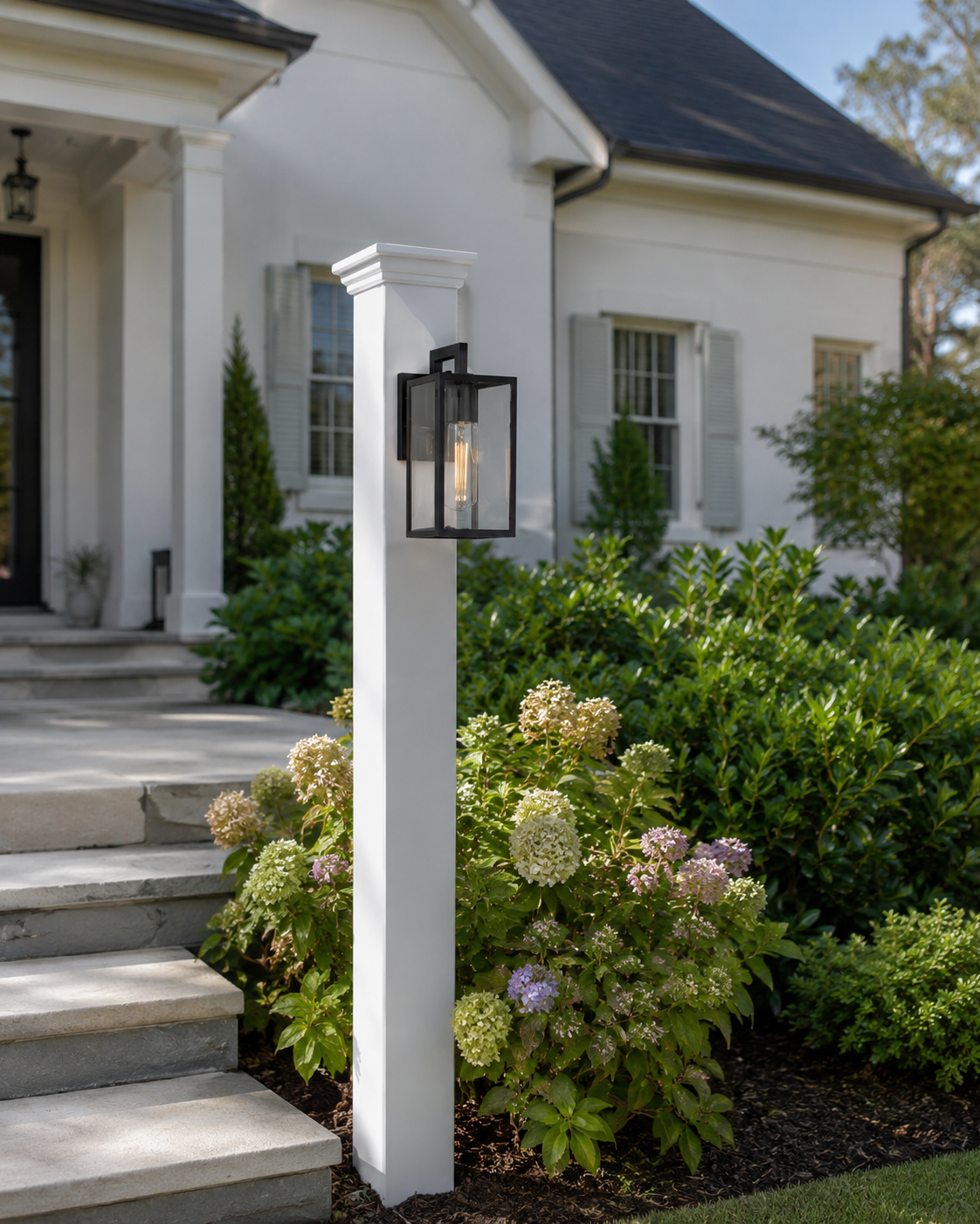 Close-up of a white porch column with a modern black outdoor wall lantern, surrounded by green bushes and flowering plants, in front of a white house with shuttered windows.