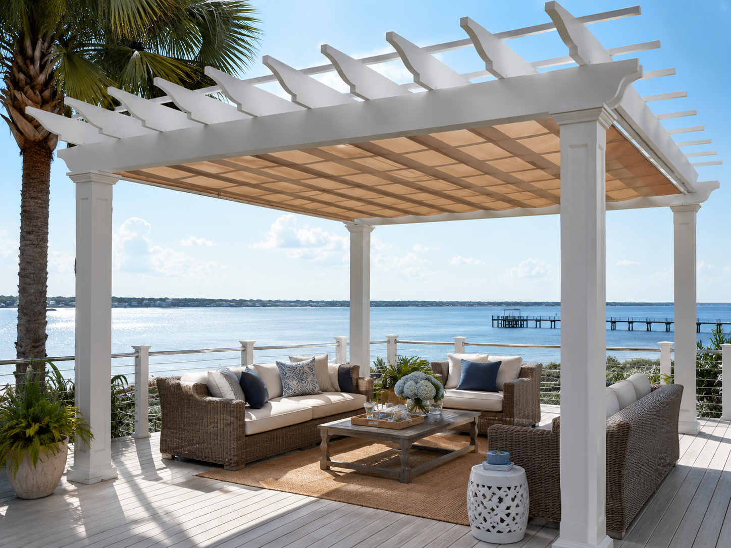 Outdoor seating area on a patio overlooking a lake, with wicker sofas, a coffee table with a floral centerpiece, and a white pergola.