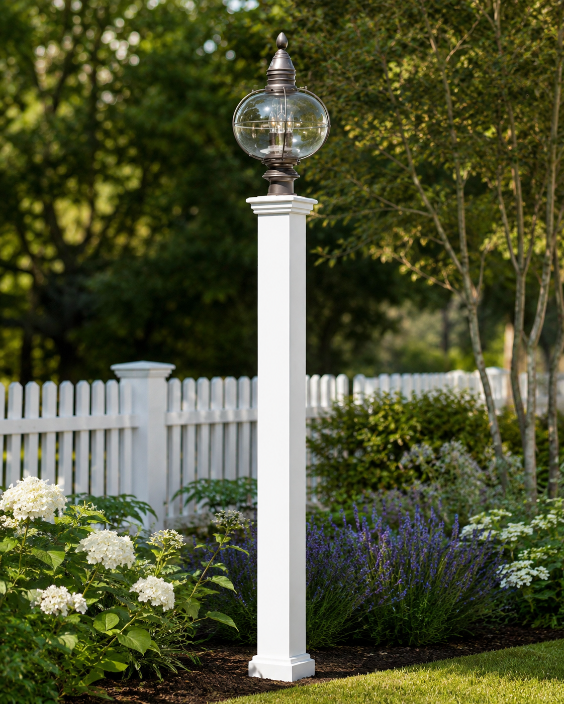 A white outdoor lamp post with a glass globe, surrounded by a white picket fence, green trees, and flowering bushes in a garden.