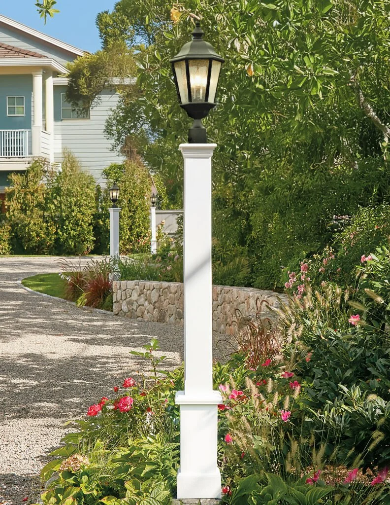 A white sidewalk lamp post with a black lantern on top, lining a gravel pathway surrounded by greenery and flowers.