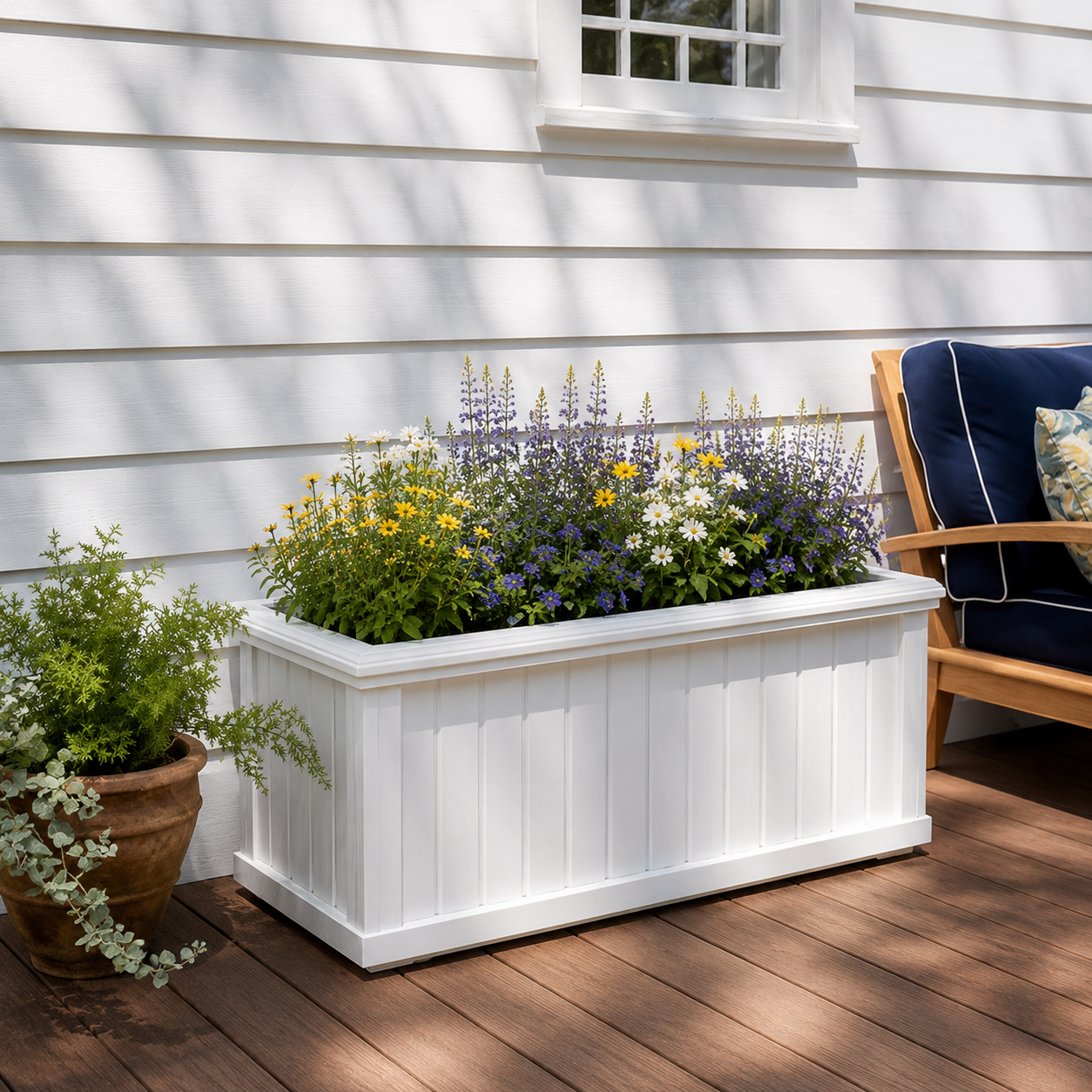 A white garden planter with colorful flowering plants, a potted green plant to the left, and a porch with wooden flooring and white siding in the background.