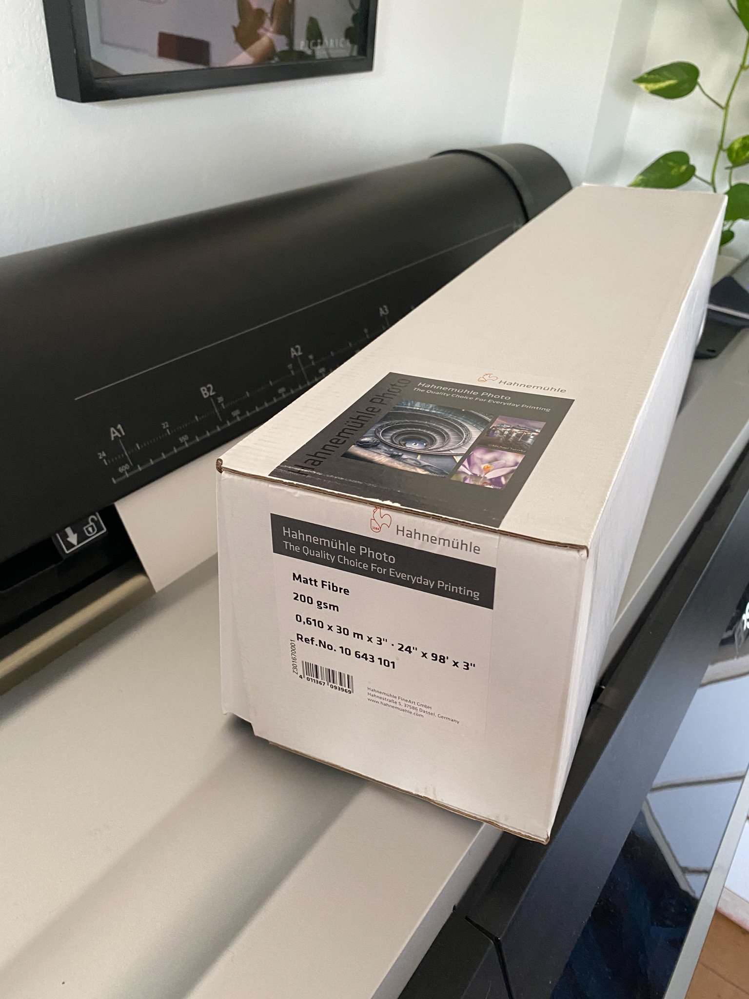 Caja de papel fotográfico Hahnemühle de color blanco sobre una mesa, cerca de una máquina de impresión y una planta en el fondo.