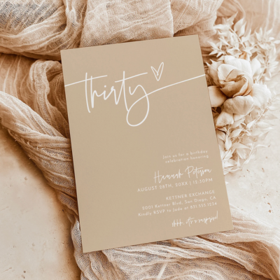 A beige invitation card with the word 'thirty' and a heart, surrounded by soft, light-colored fabrics.