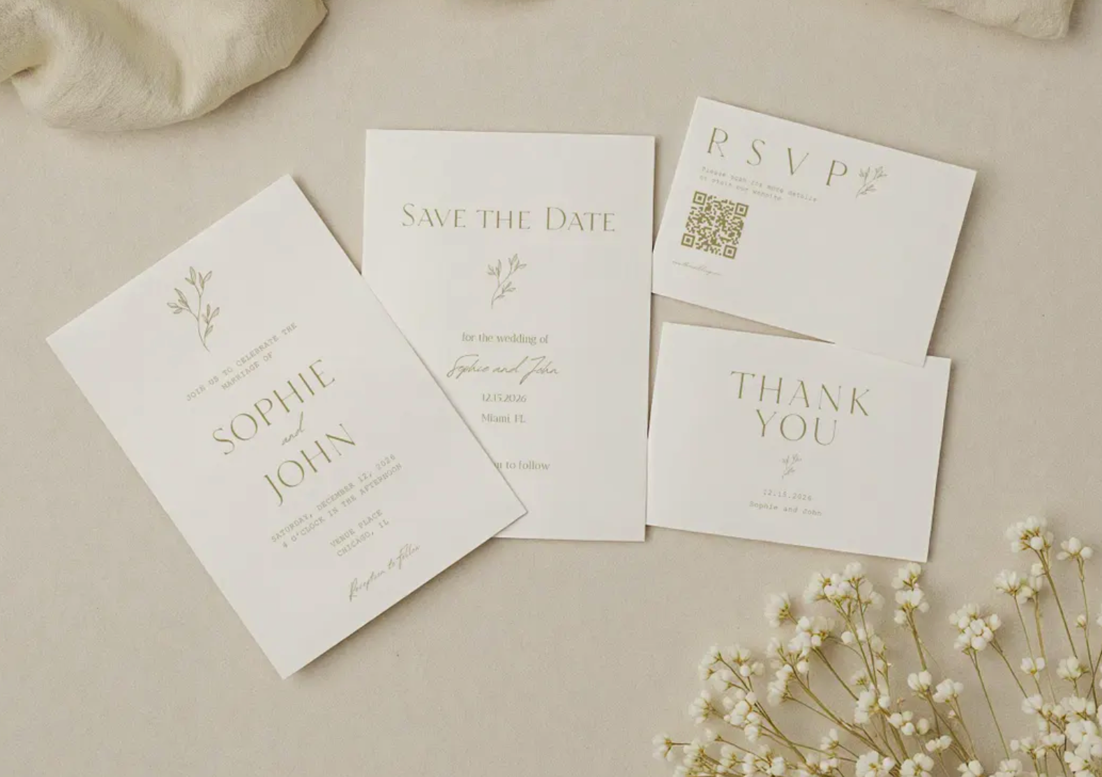 Wedding invitation stationery including a save the date card, RSVP card, and thank you card, along with white flowers on a beige surface.