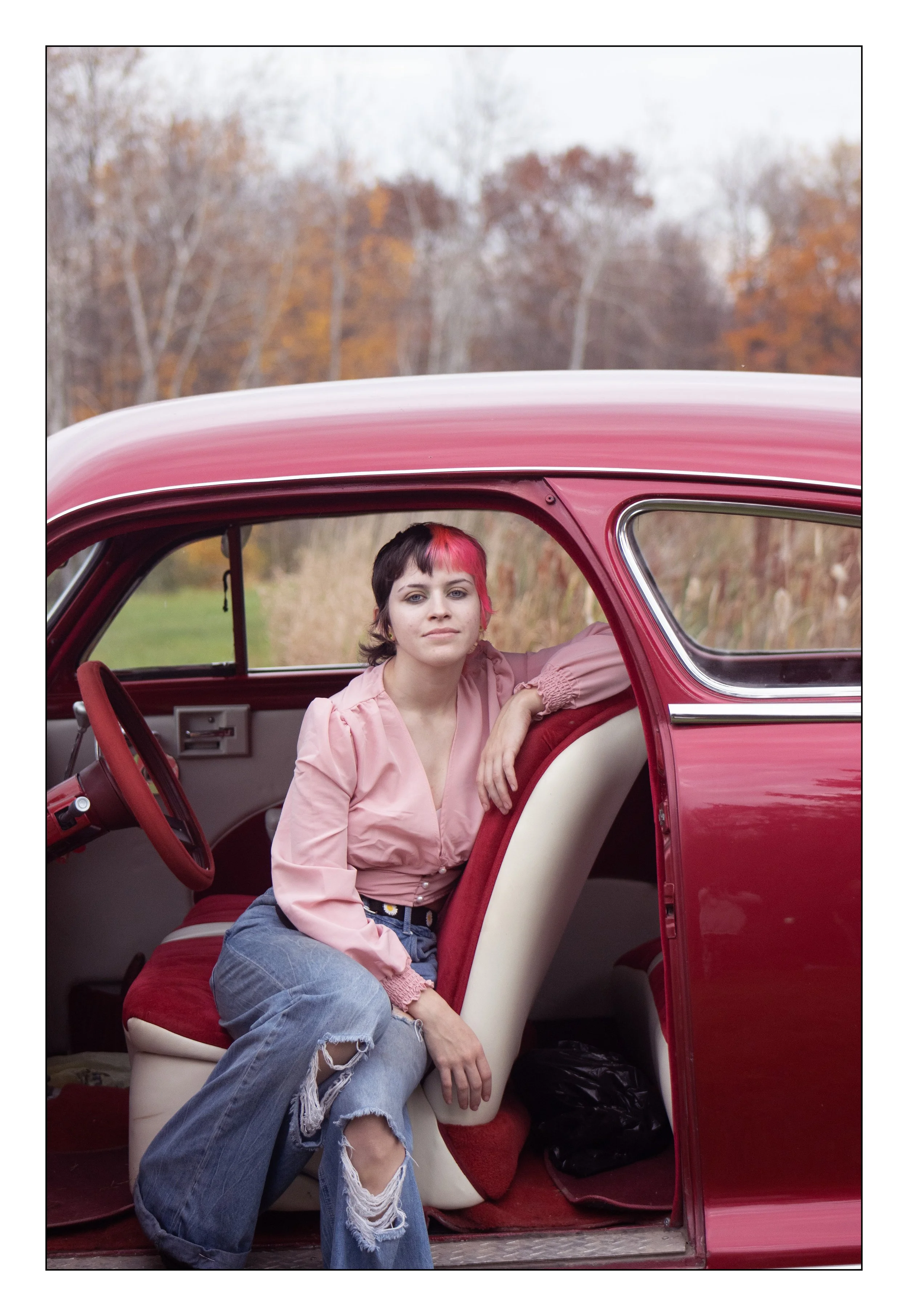 Young woman with short black and pink hair sitting inside a vintage red car, outdoors during autumn with trees in the background.