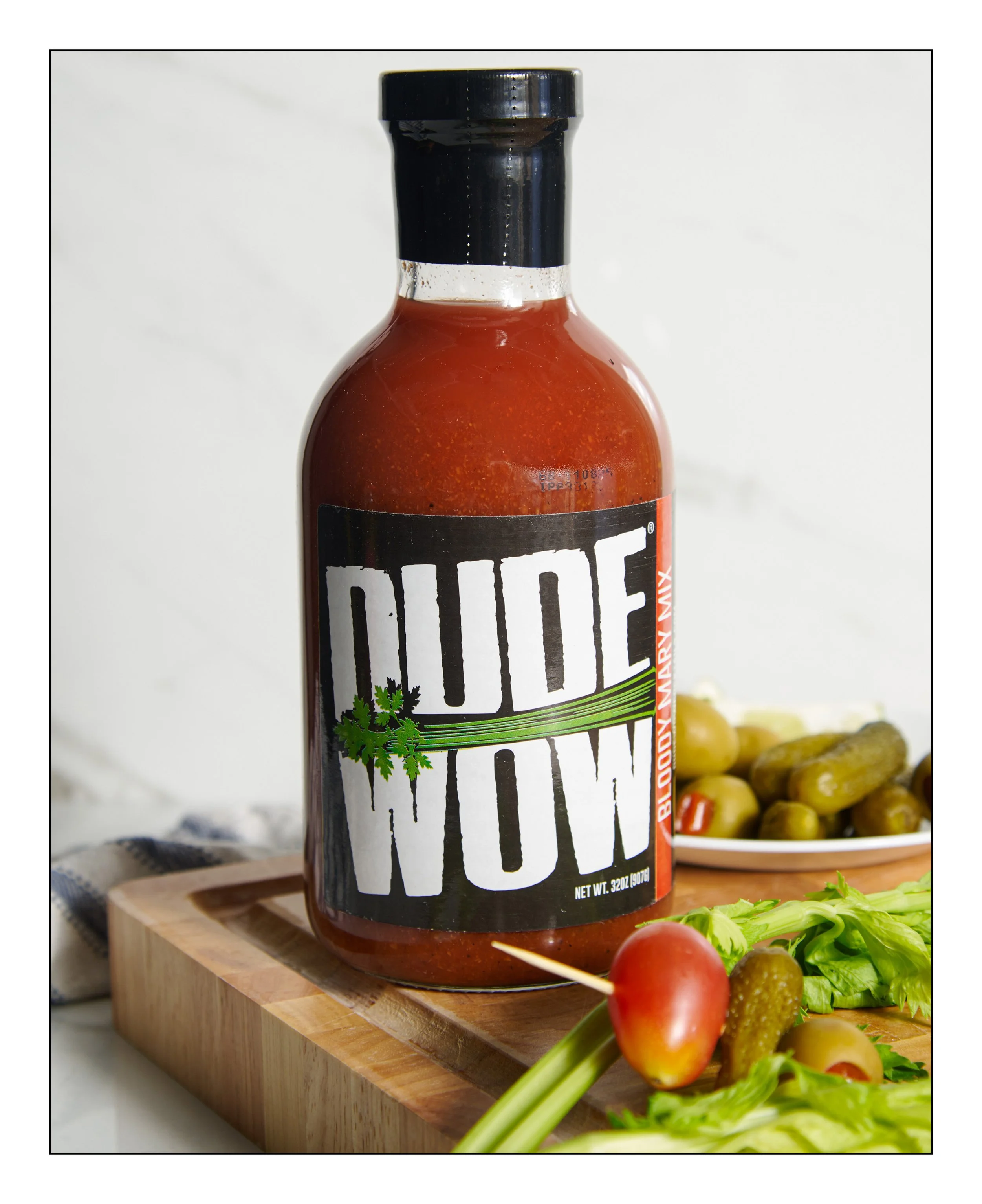 A bottle of Dude Wipes Bloody Mary Mix on a wooden cutting board with celery, cherry tomato, pickles, and lettuce in the background.