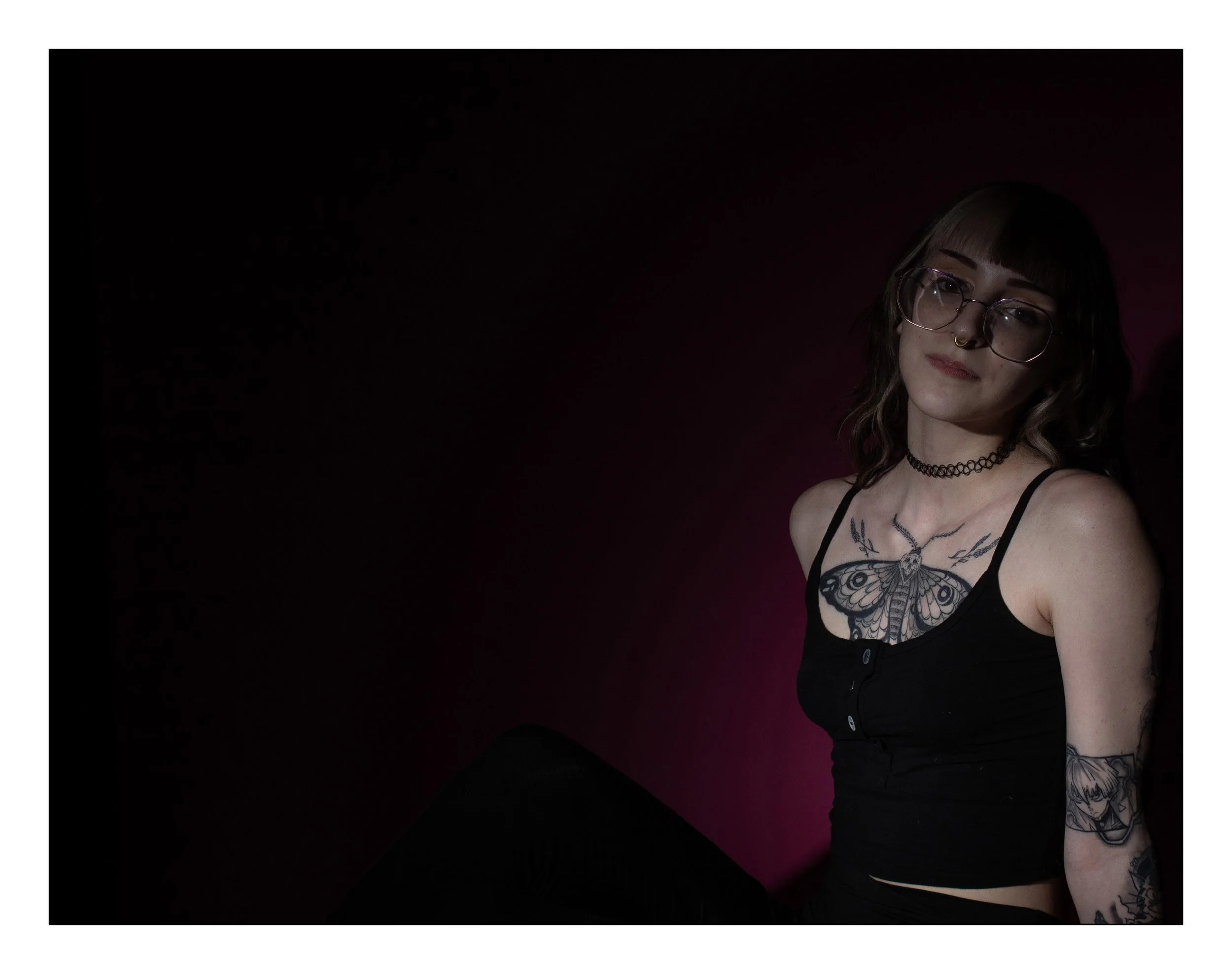 Young woman with glasses, tattoos, and a choker necklace seated against a dark background with pink highlights, wearing a black tank top.