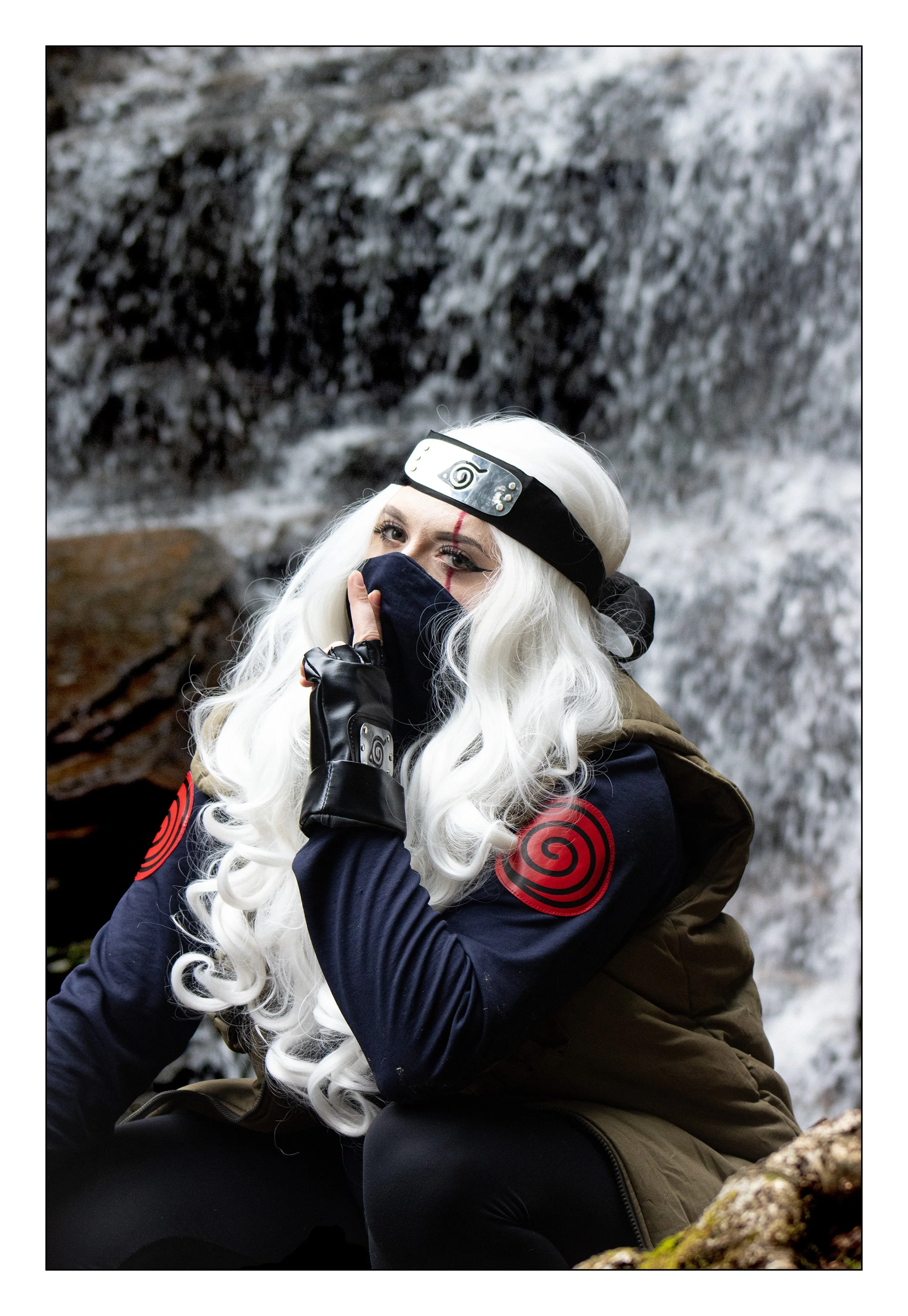 A cosplayer dressed as Kakashi from Naruto, wearing a black face covering, a Naruto headband, black gloves, and a dark jacket with red spiral symbols, sitting near a waterfall background.