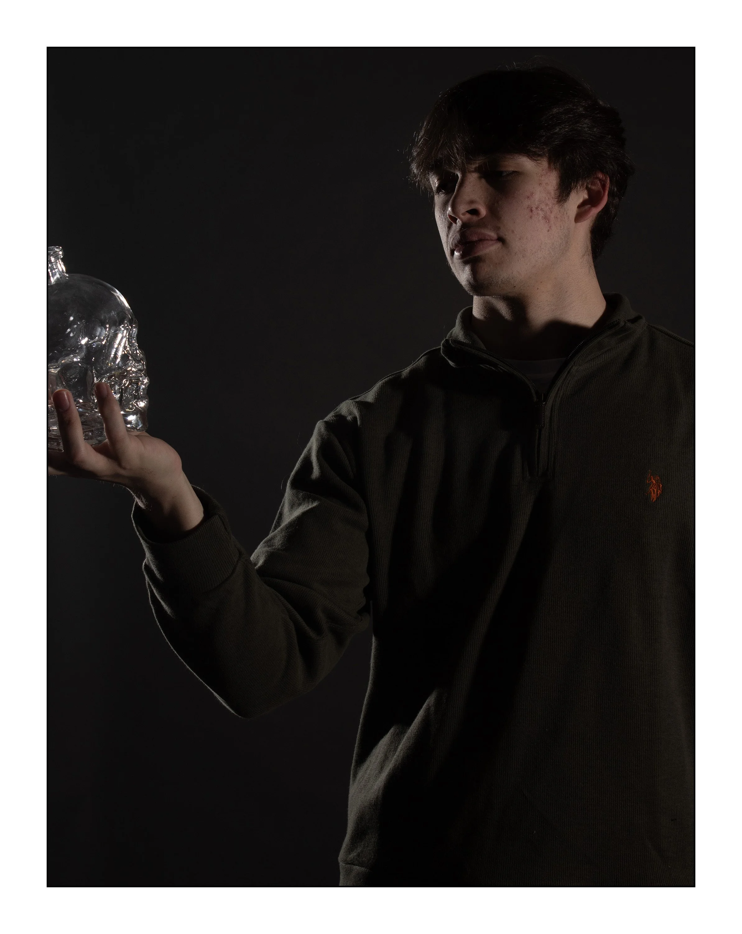 A young man holding a clear, glass skull sculpture against a dark background.
