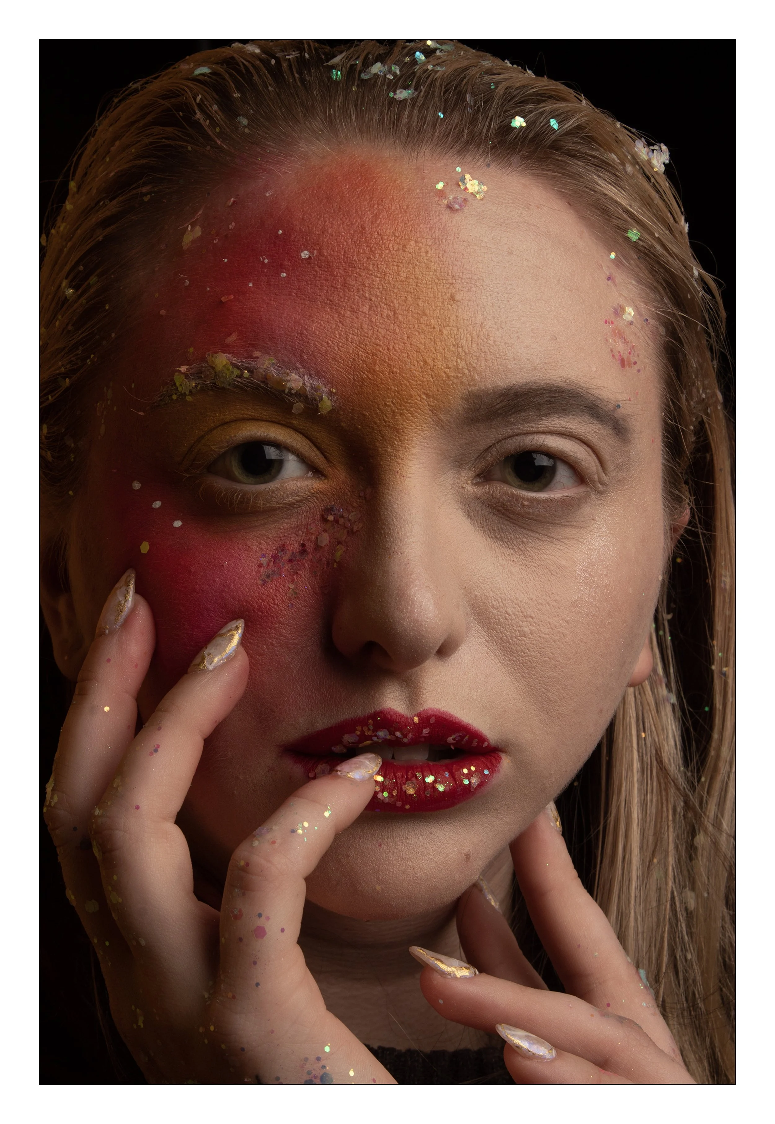 A close-up portrait of a woman with half of her face painted in vibrant colors and decorated with glitter, while the other half remains natural. She has intense gaze, red lipstick, and her fingers adorned with glitter nail polish.