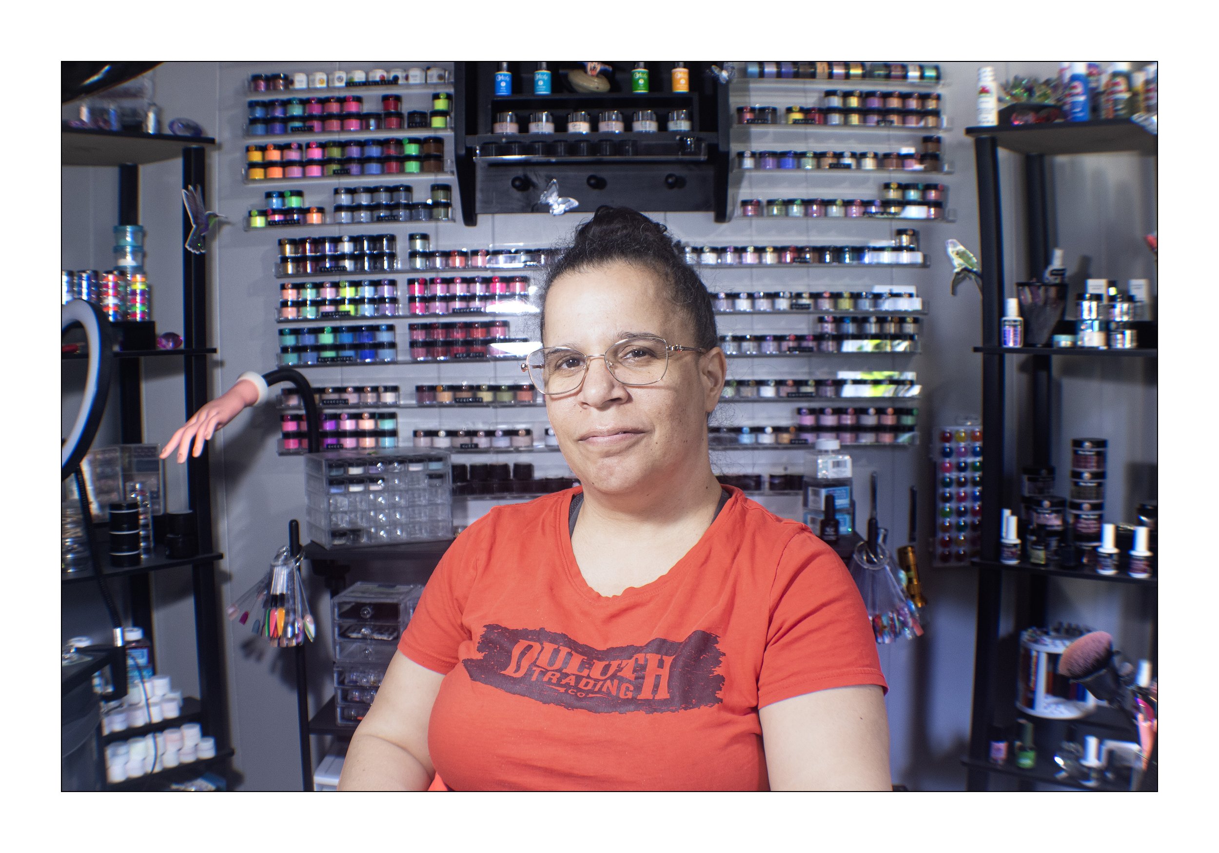 A woman with glasses and dark hair in a bun wearing an orange T-shirt sitting in a colorful arts and crafts supply store with shelves of glitter, paints, and craft supplies.