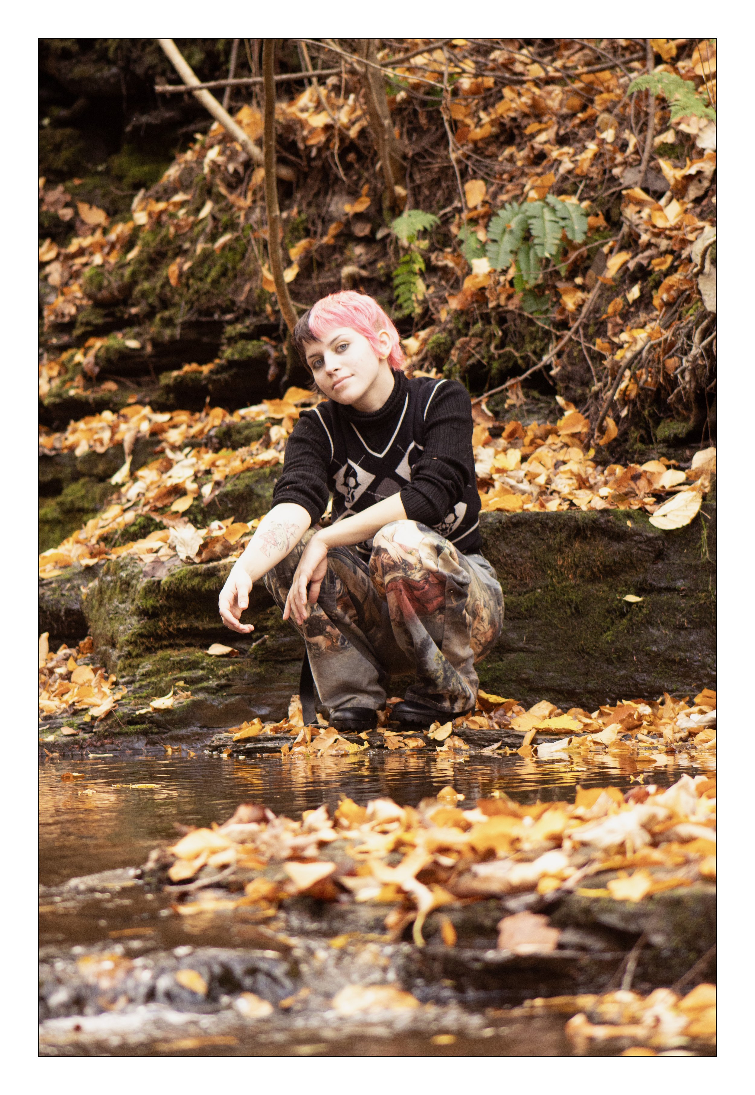 A young person with pink hair sitting by a small stream surrounded by autumn leaves in a forest.