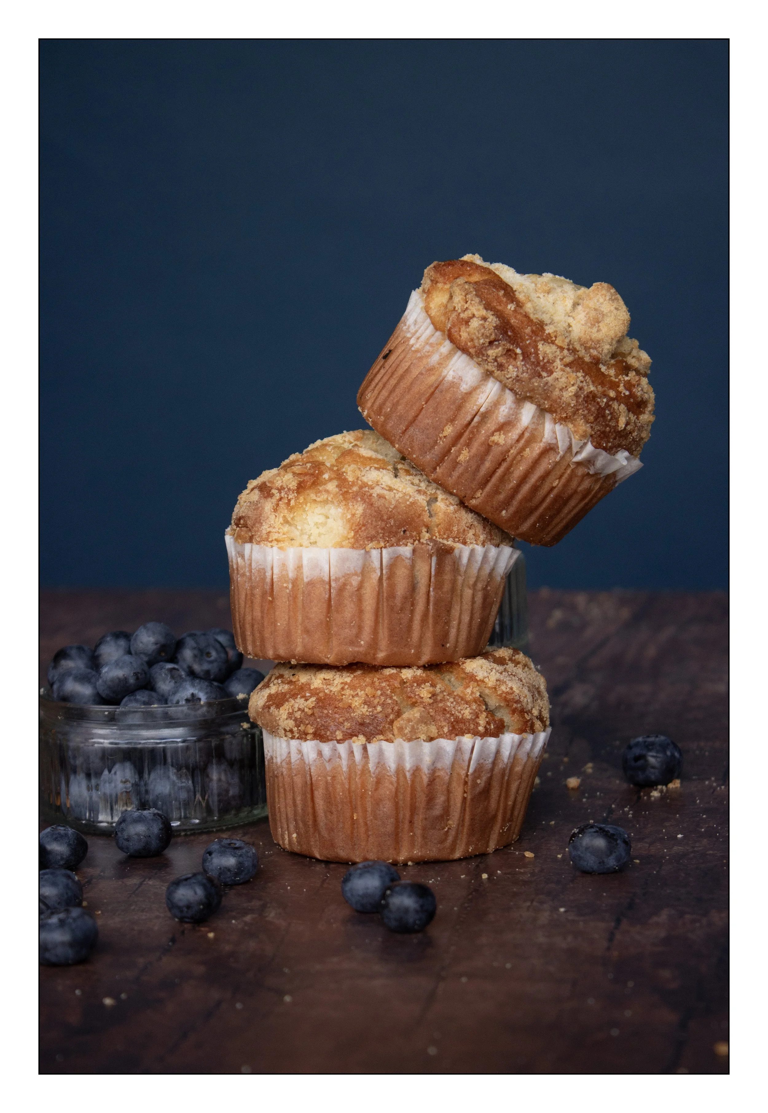 Stacked blueberry muffins on a dark wooden surface with blueberries in a small glass bowl beside them, against a dark blue background.