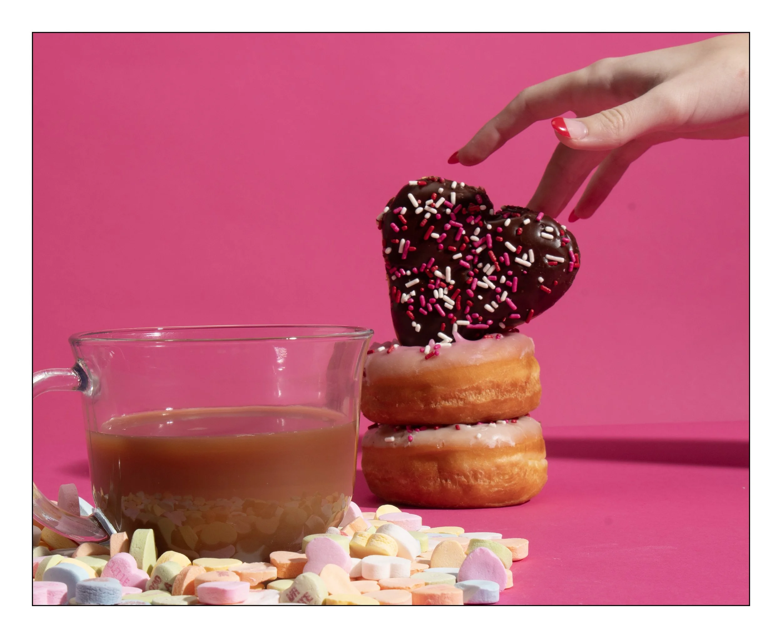 A hand with red nail polish reaching for a heart-shaped chocolate donut with pink, white, and red sprinkles on top, stacked on two glazed donuts, next to a clear mug of tea on a pink surface surrounded by colorful marshmallows.