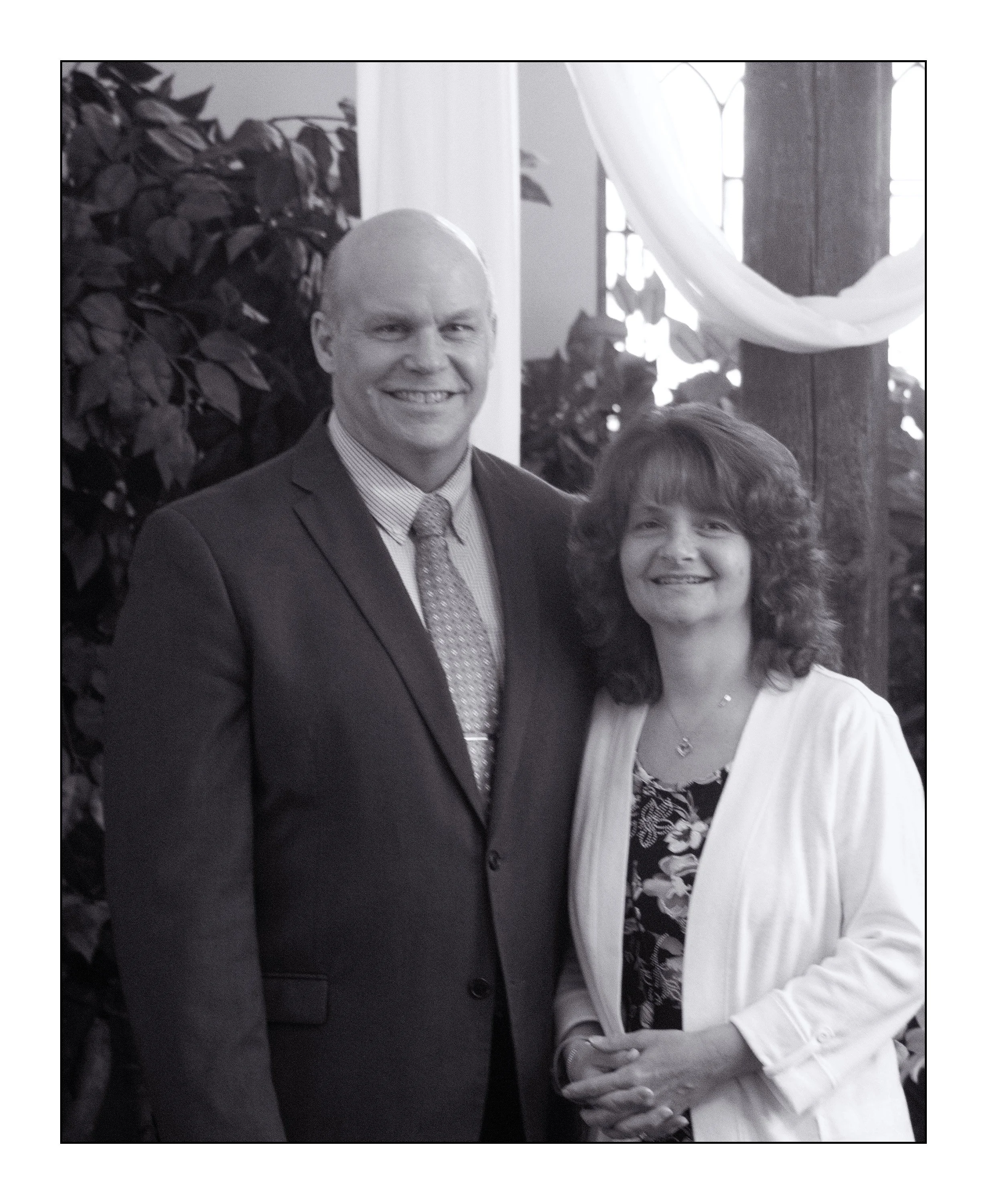 A black-and-white photo of a man in a suit and a woman in a blouse and cardigan standing close together indoors, smiling. Behind them are large plants, windows, and draped fabric.