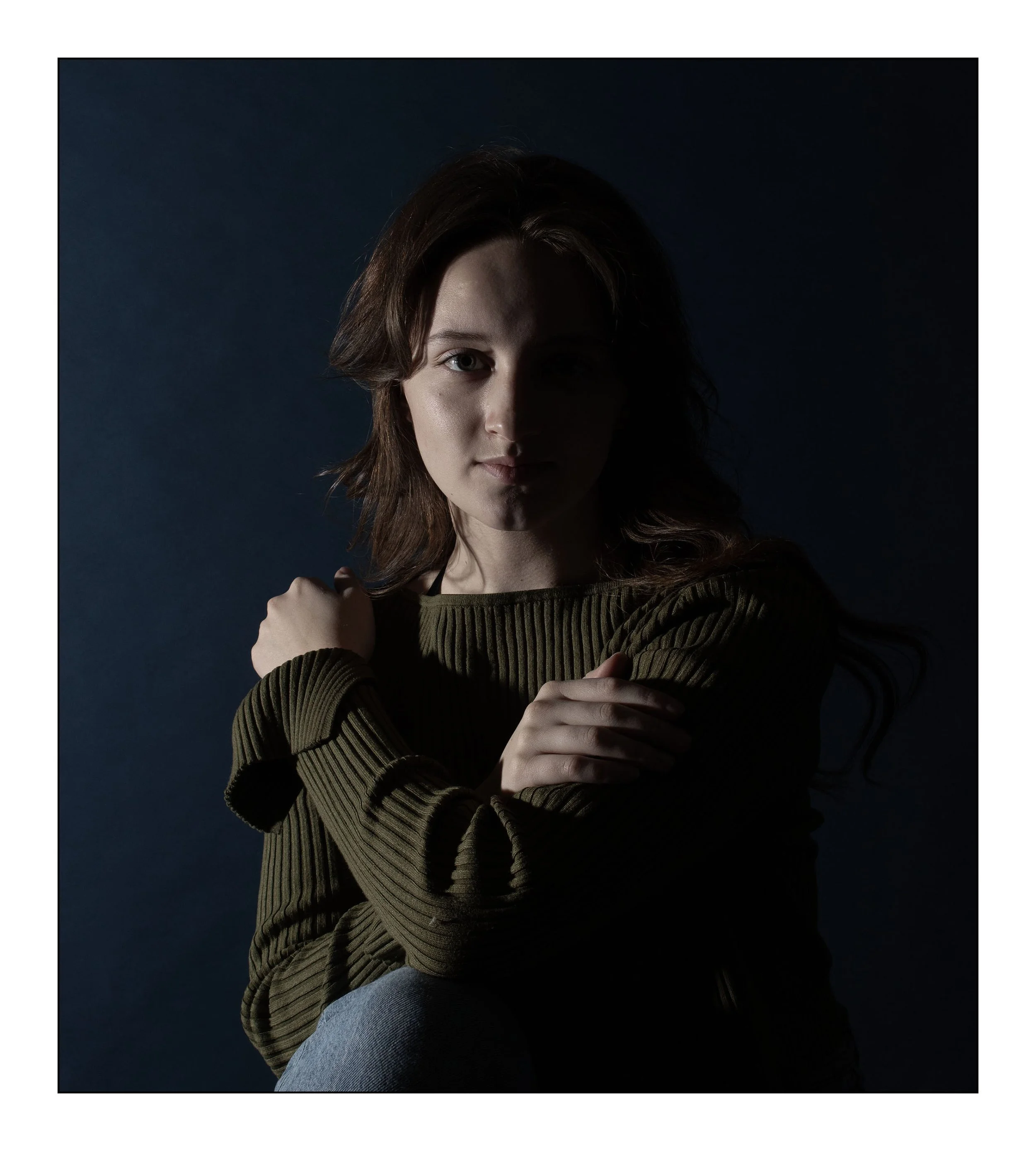 Portrait of a young woman with brown hair, dark background, wearing a green ribbed sweater, crossing her arms and looking at the camera.