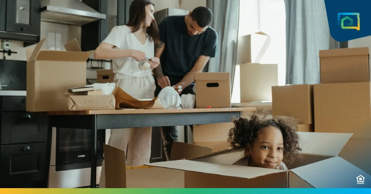 Man and woman unpacking boxes and a child in the forefront peeking out of a cardboard box