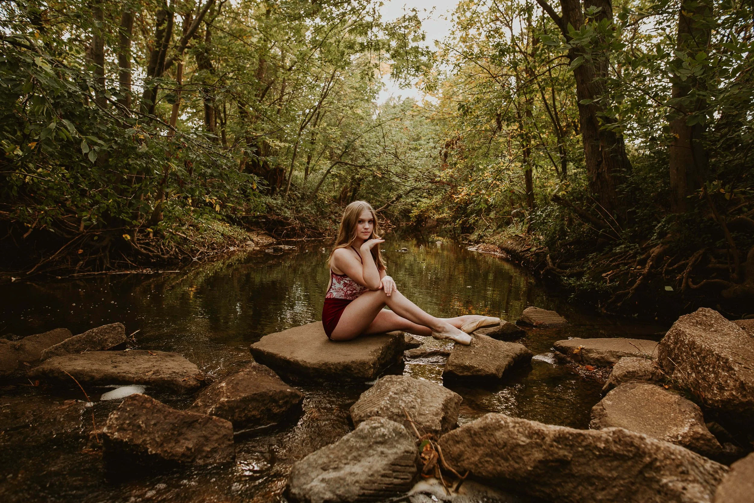 High school dancer sits atop a large rock with her toes pointed, chin resting atop her hand.