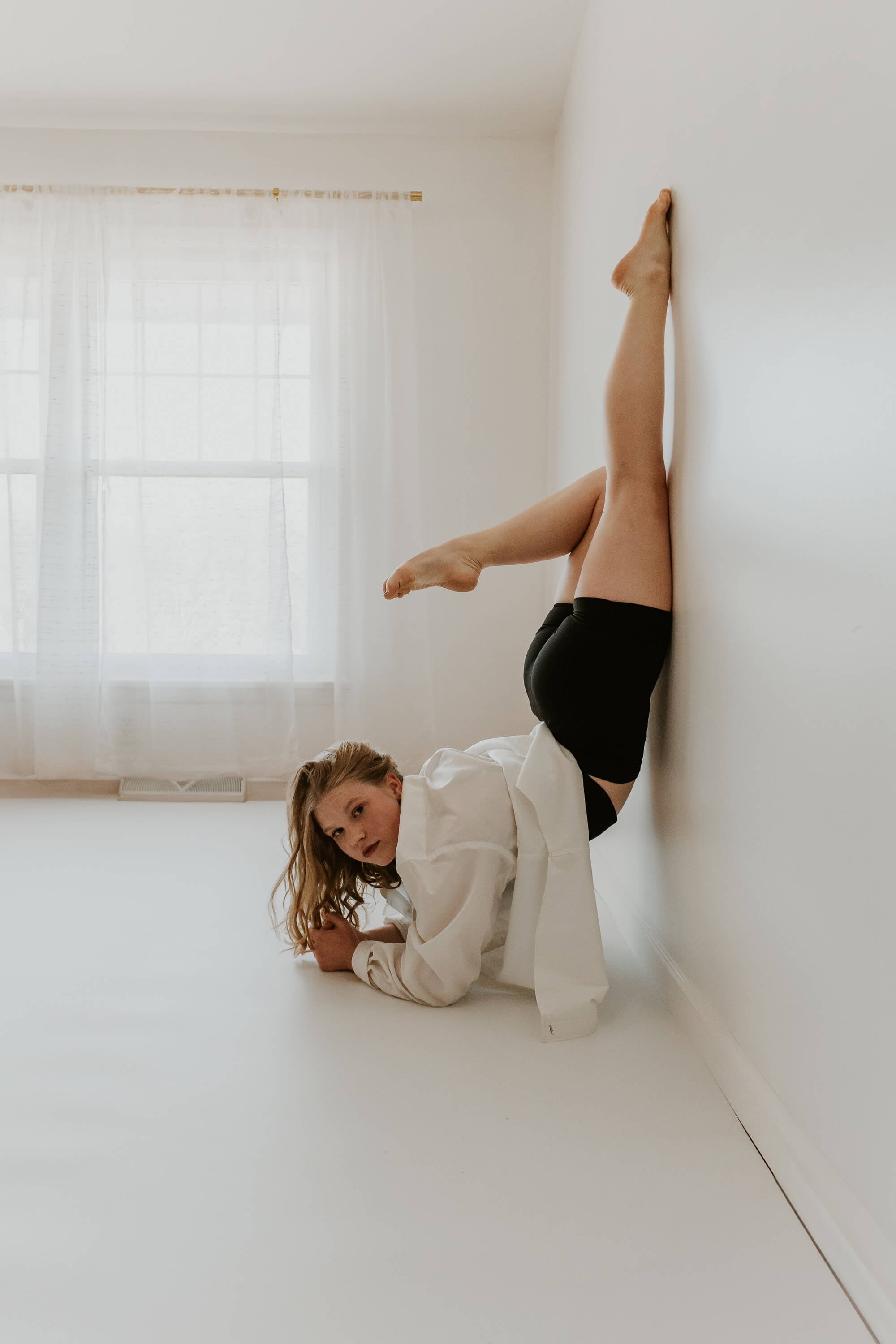 A dancer wearing a white dress shirt smles as her body rests vertically against a white wall.