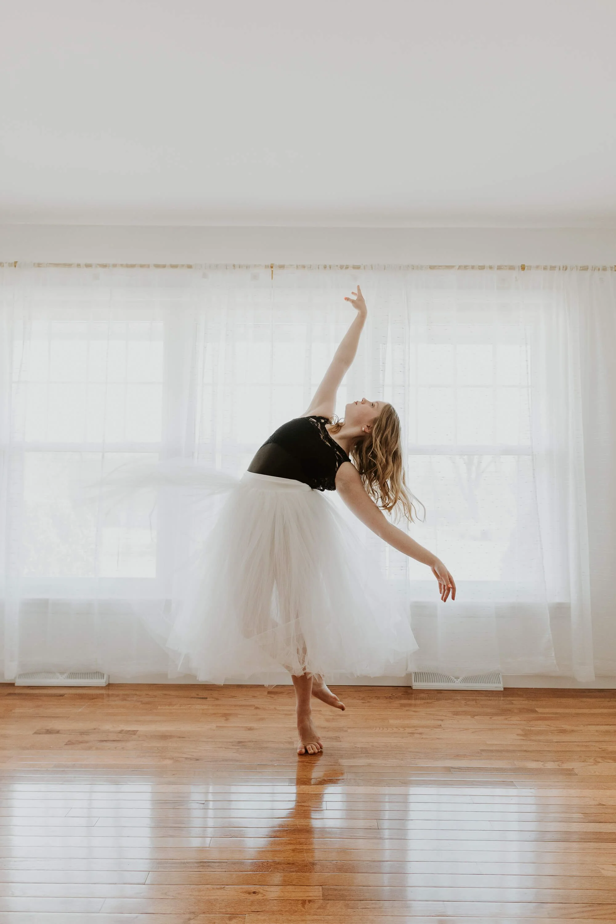 A dancer wearing a romatic, white tutu and black leotard holds a graceful pose in front of a wall of windows.