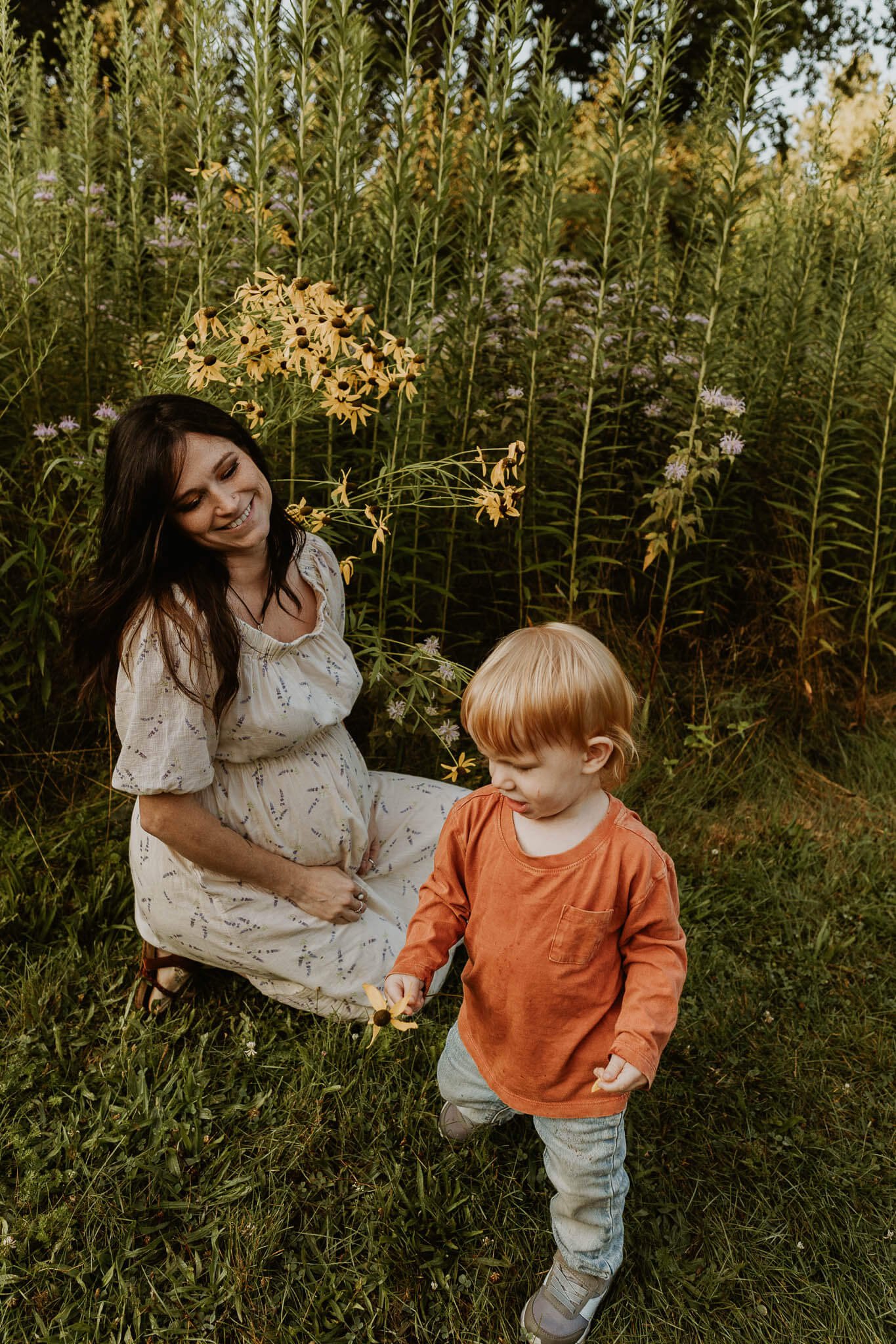 A mom expecting her second child smiles at her first born as he navigates a field of prairie flowers.