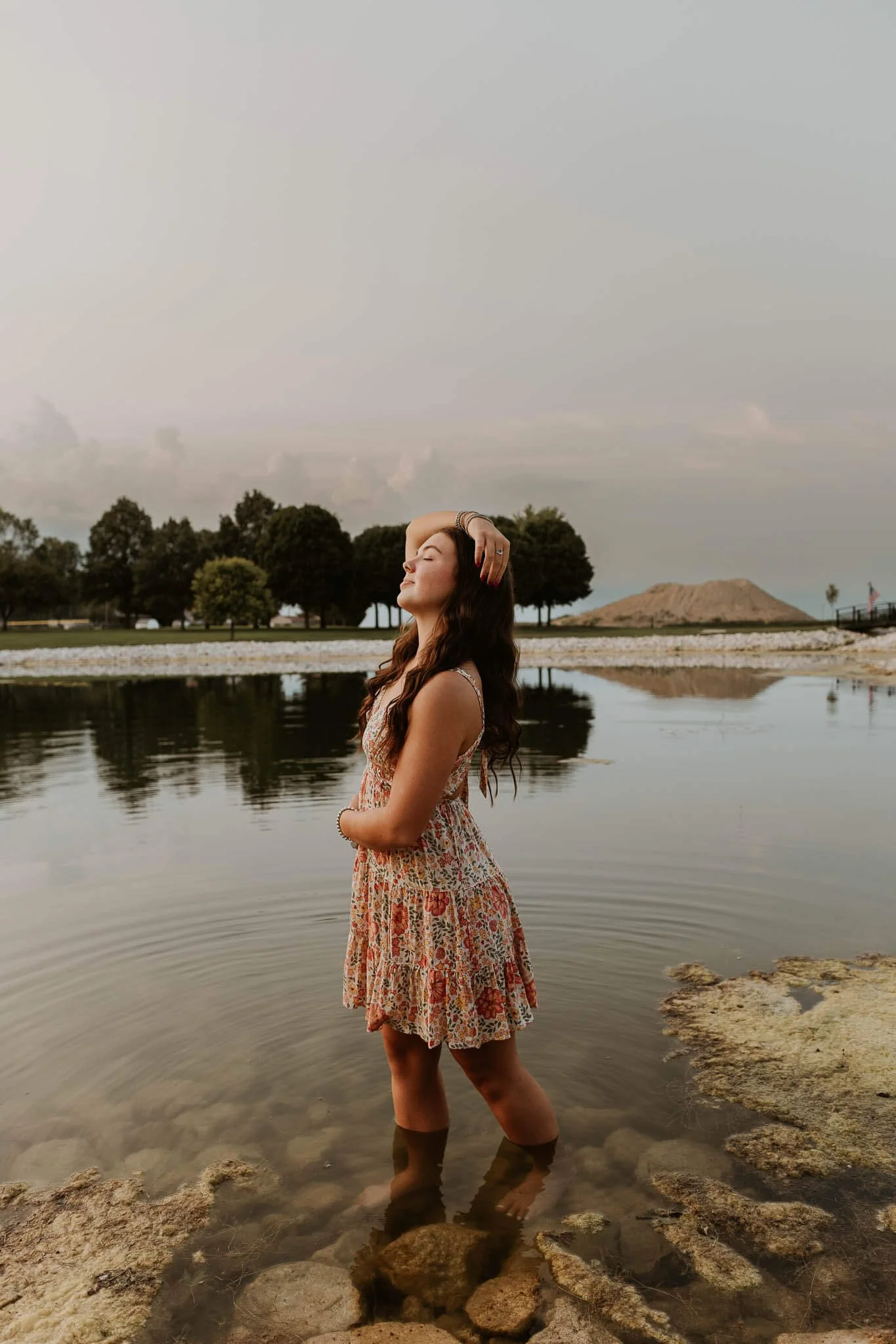 A senior from Olympia High School holds a pose with one arm wrapped around her waist and the other over her head, in a pond at Hopedale Park.