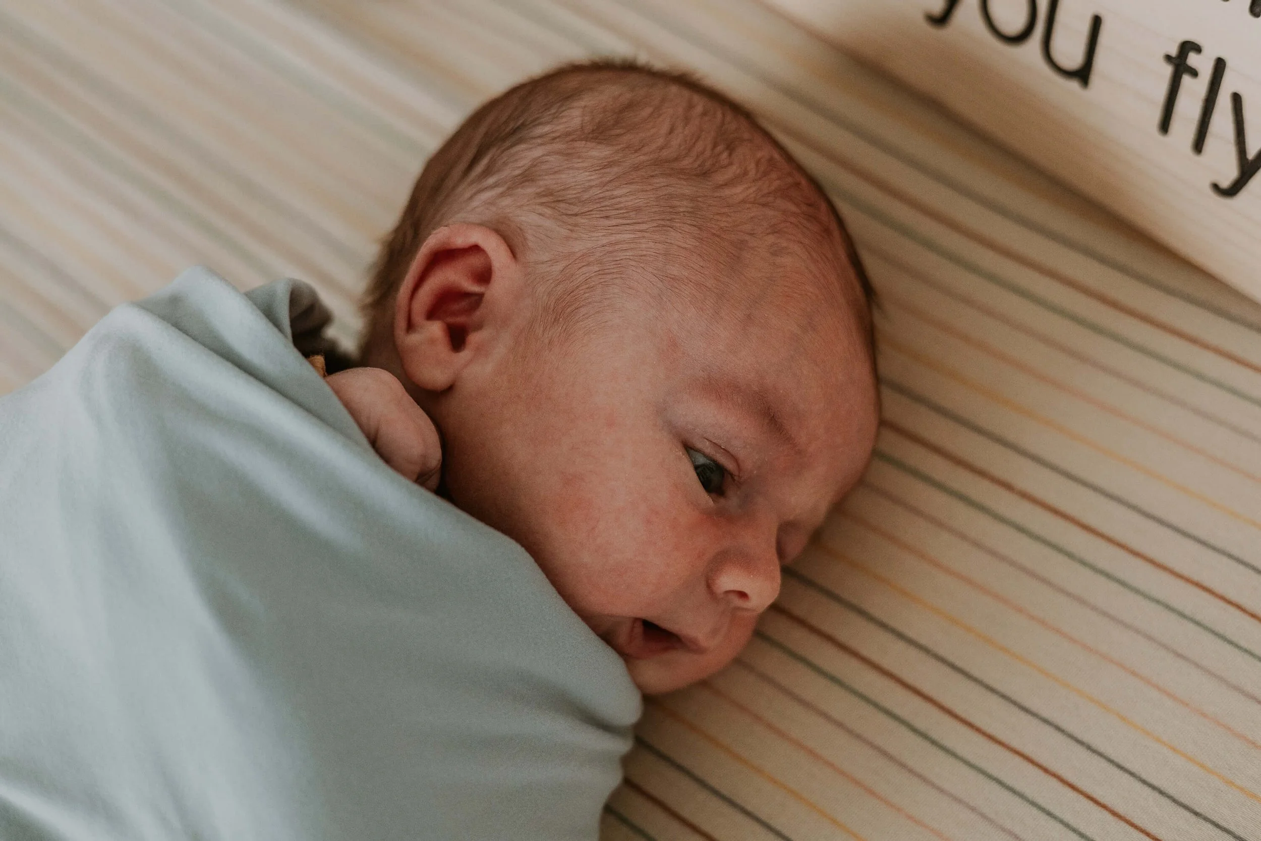 A newborn swaddled in a blue blanket, eyes wide open and head turned to the side