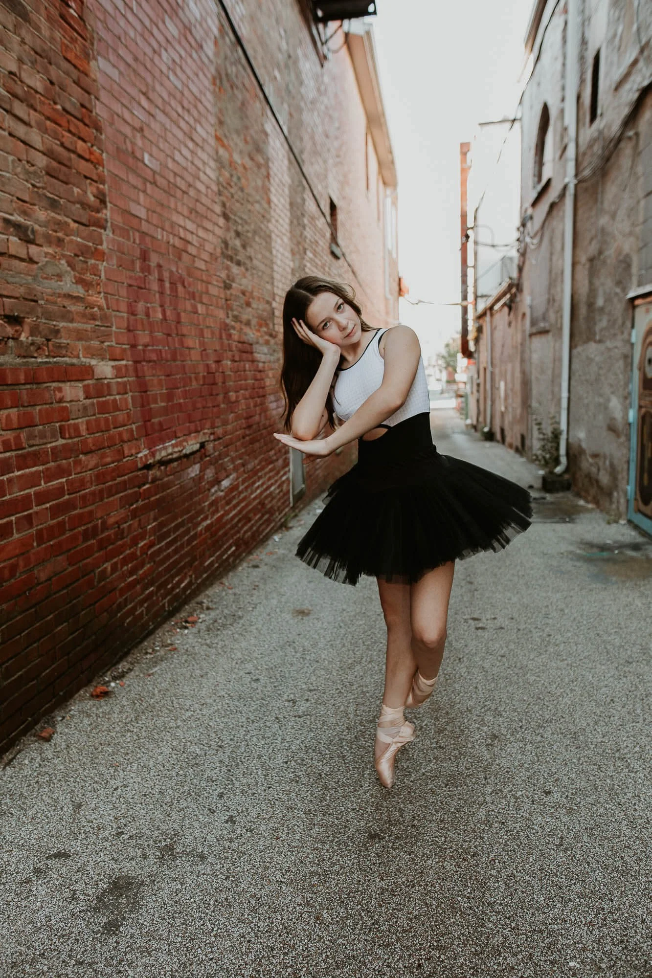 A dancer in an alleyway casually poses with her elbow in hand, check resting in her palm.