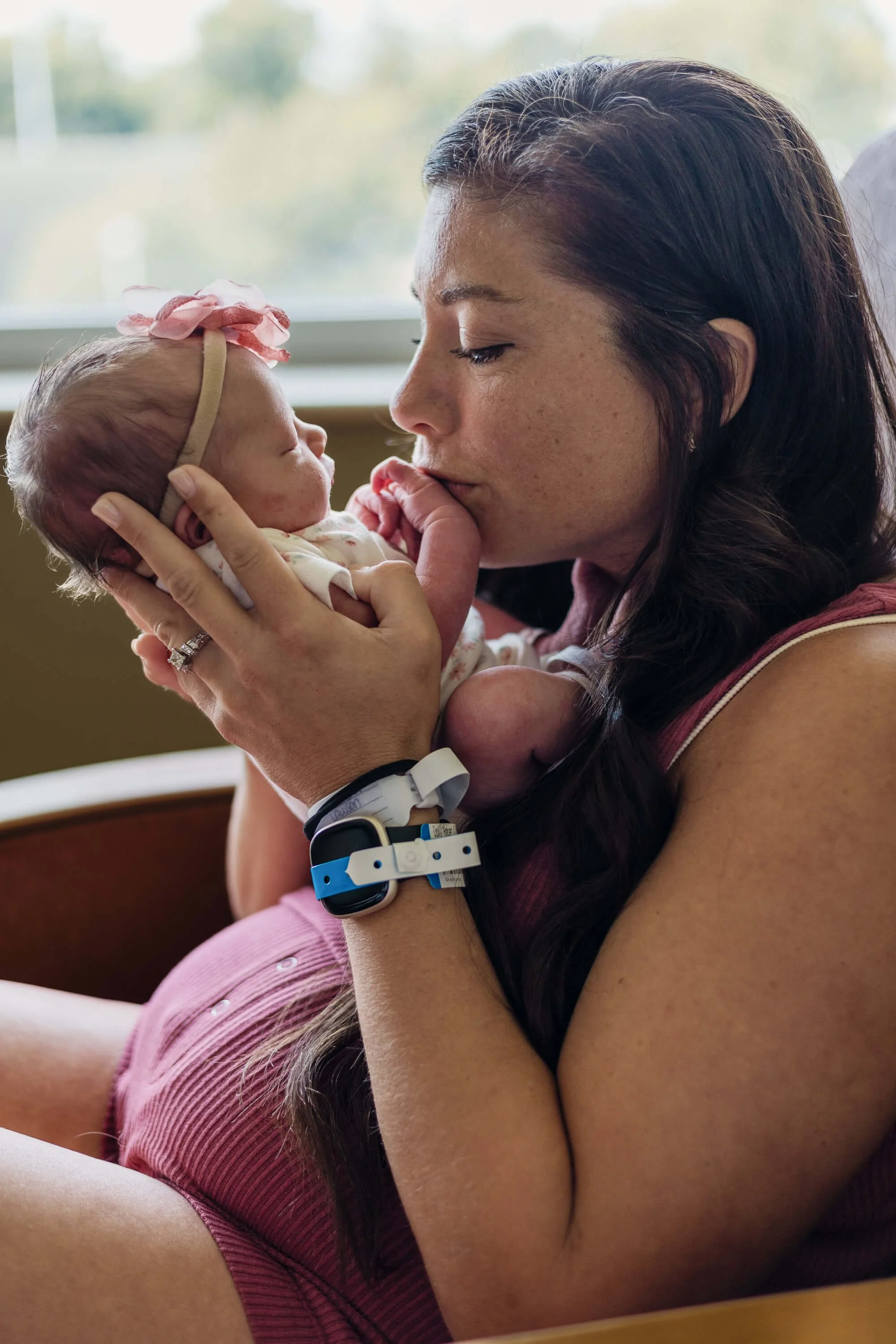 A newly postpartum mother kisses her newborn daughter's fingers while sitting in a rocking chair next to her hospital bed