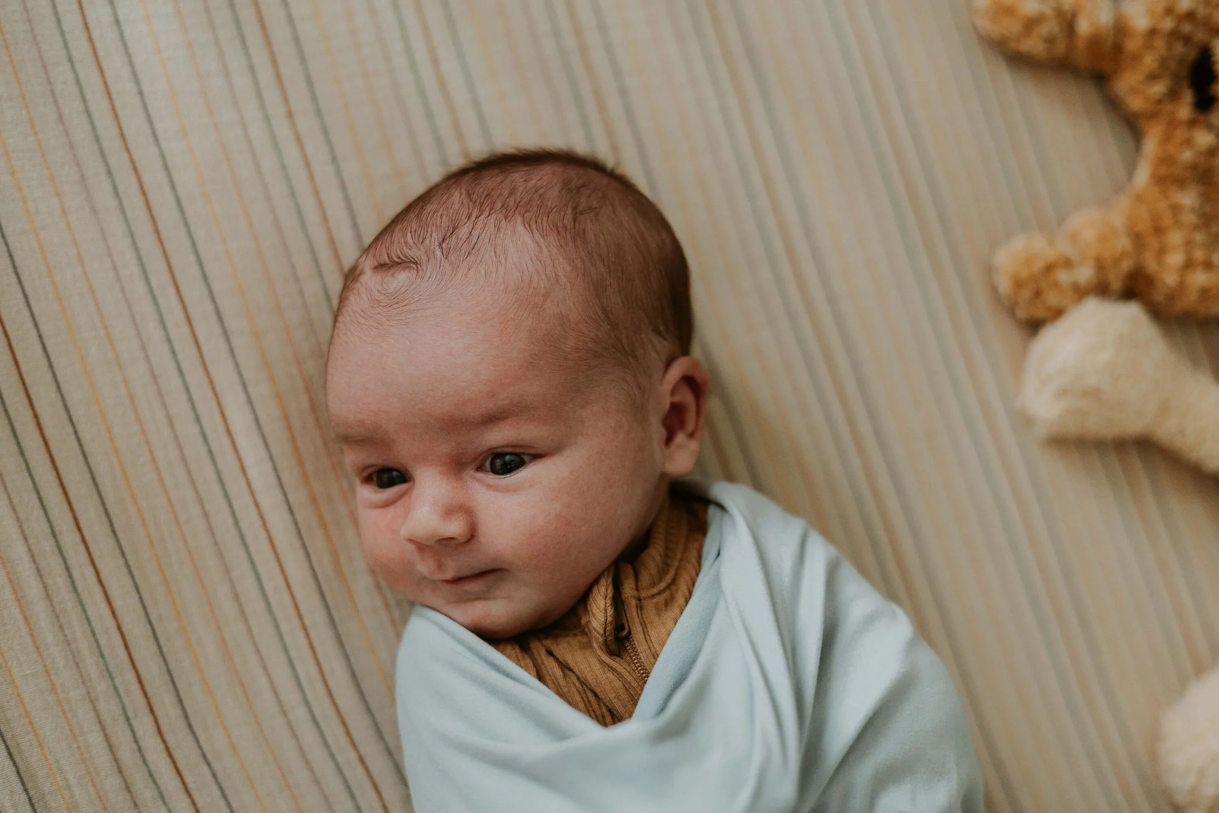 A close up of a newborn boy who looks as though he has the tiniest hint of a smile while he lays wide-awake in his crib.