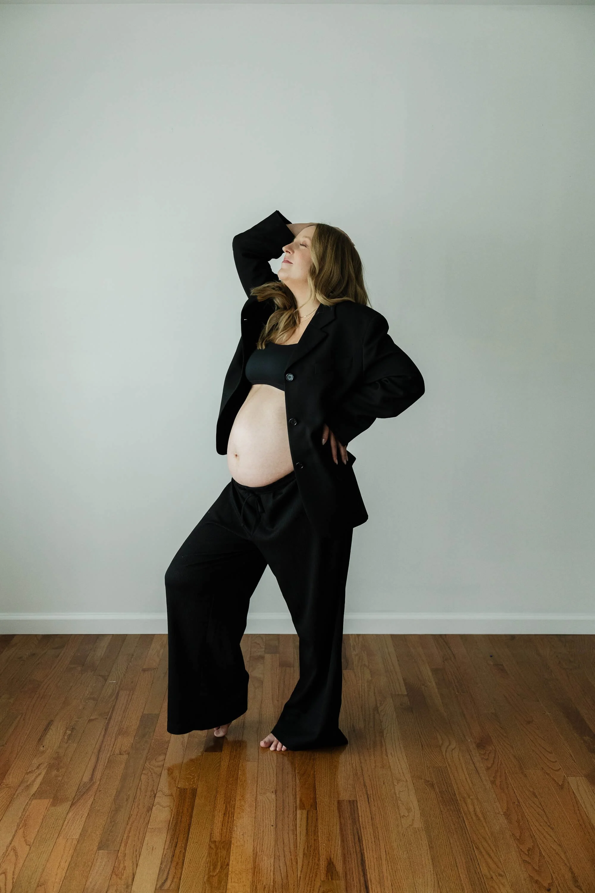 A pregnant woman dressed in black dress pants, black blazer, and black tube top strikes a pose during her maternity session in Bloomington IL