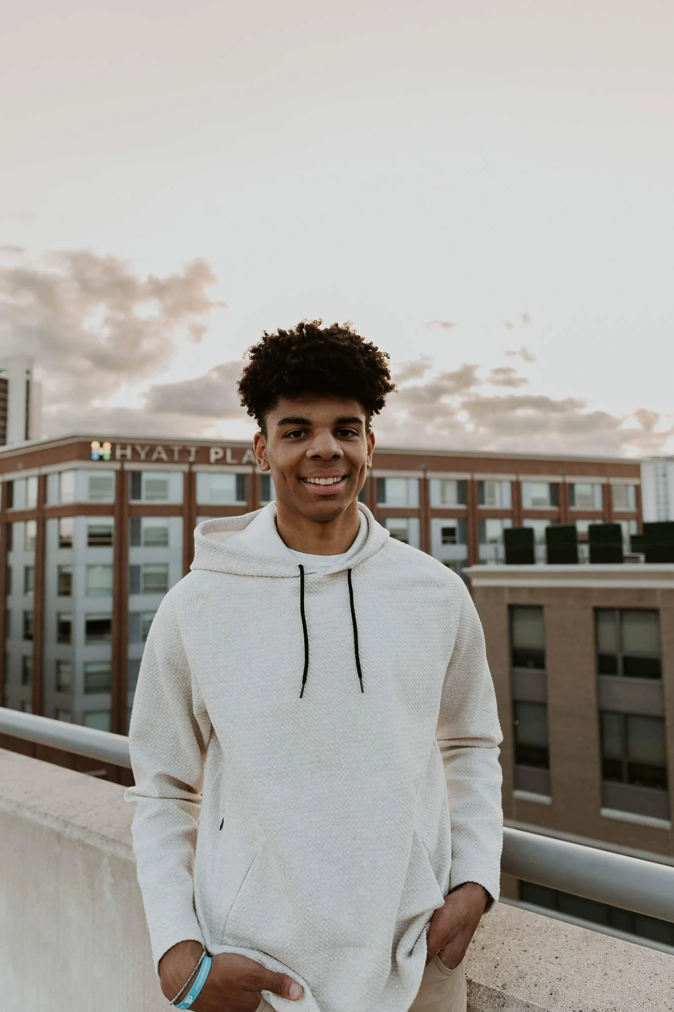 A teen boy smiles with his hands in his pockets, The Hyatt Place in Normal, IL seen in the background.