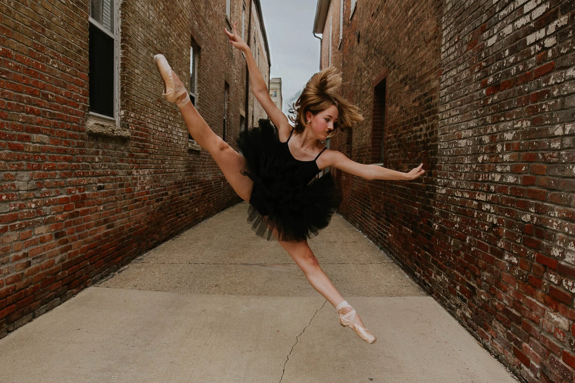 A ballerina dressed in a black tutu and leotard leaps up in a split pose, showing her strength and grace in an alleyway