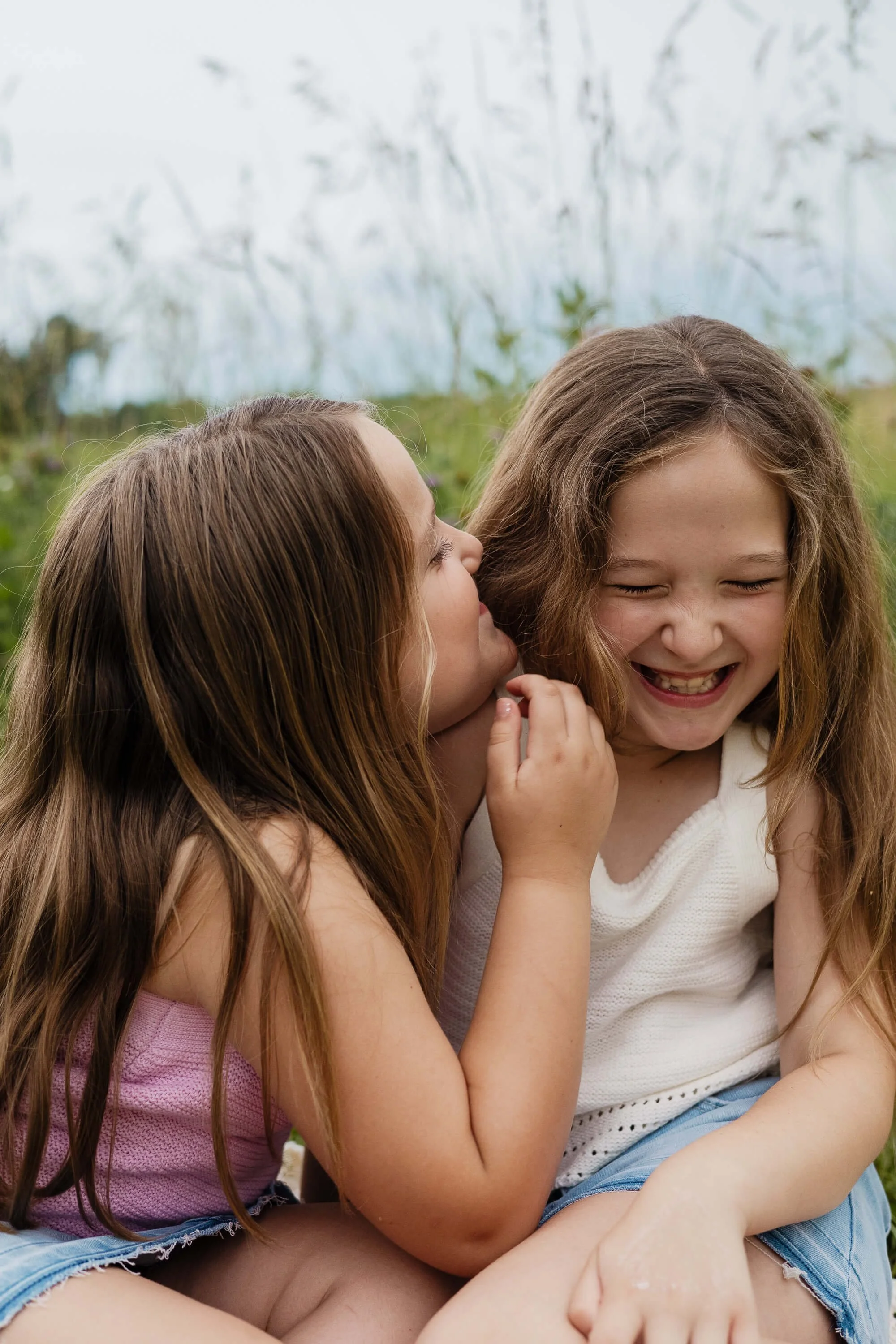 A young girl giggles as her sister whispers playfully into her ear