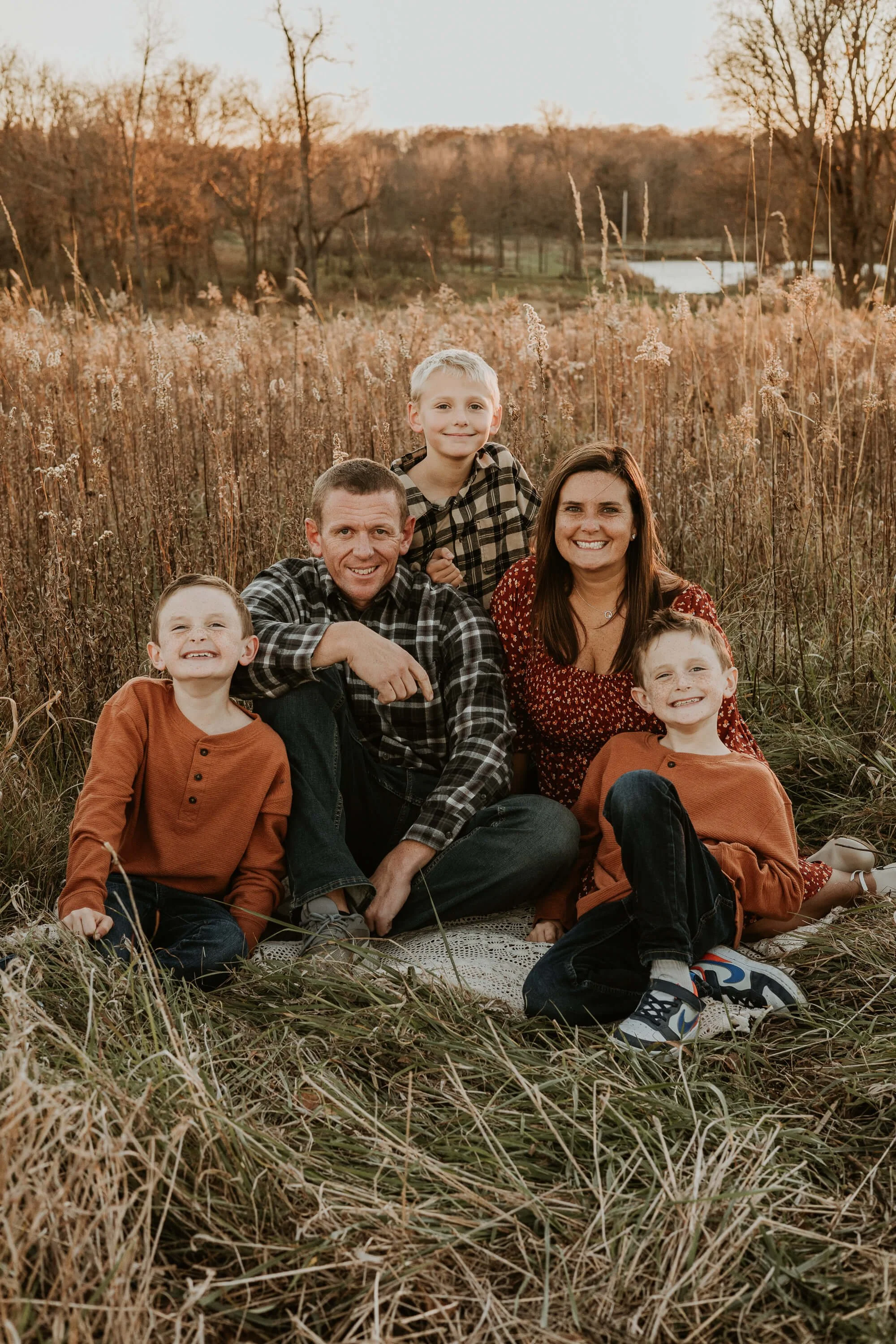 Parents and their three young boys sit cozily on a blanket in a field that overlooks a pond