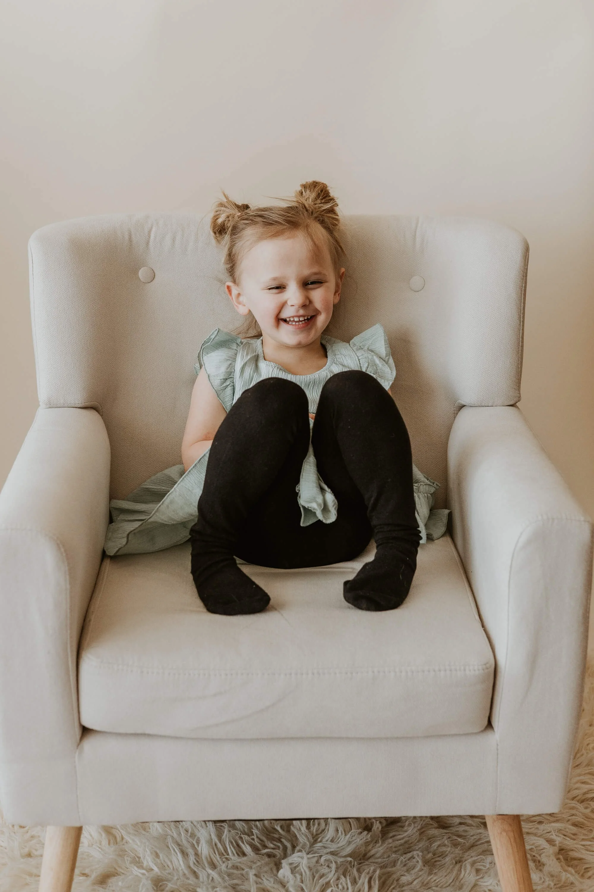 A three year old girl crinkles her nose in a laugh, sitting up in a cream chair