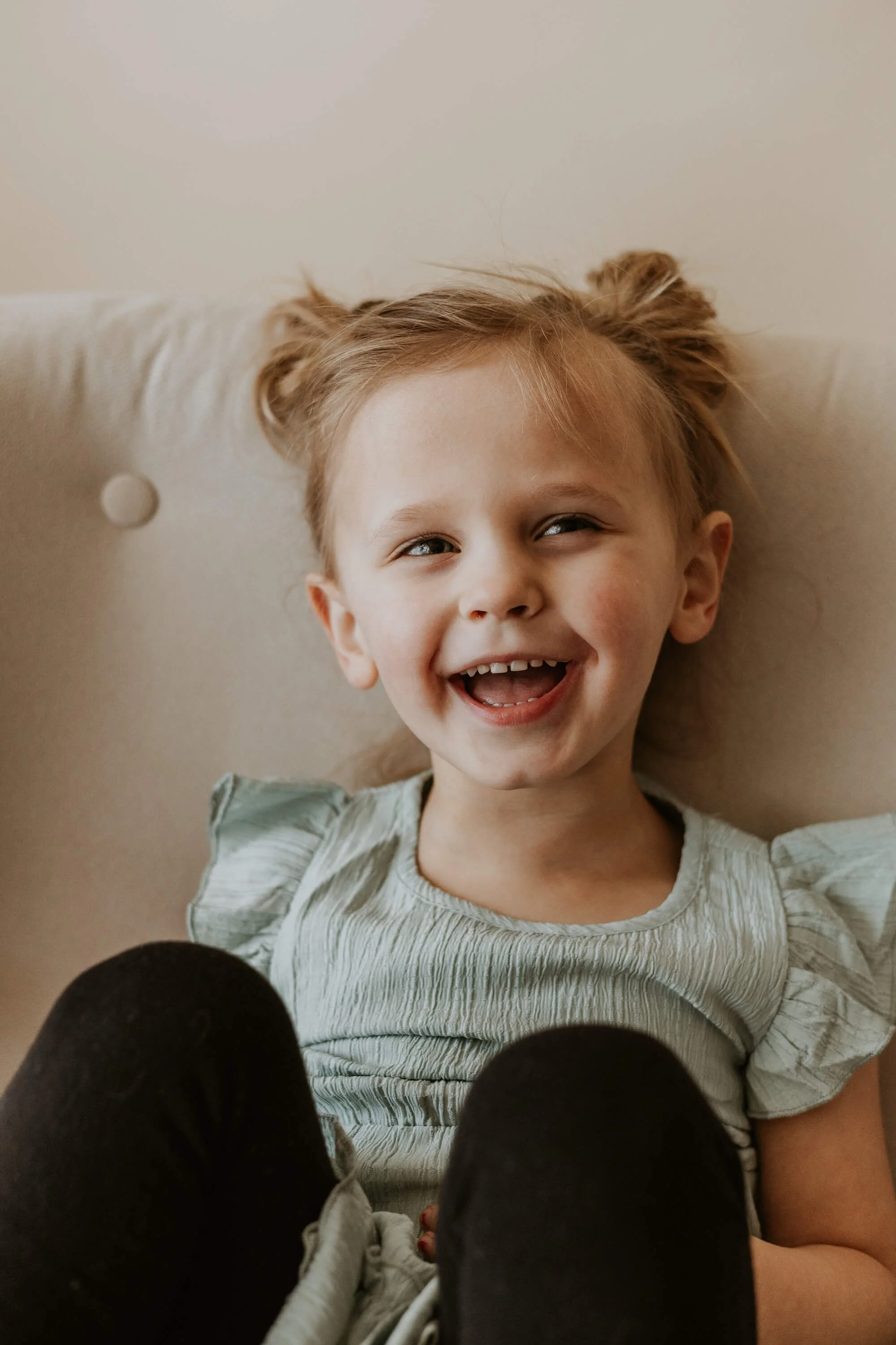 A 3 year old laughs playfully while sitting in a cream chair in a studio in Bloomington, IL