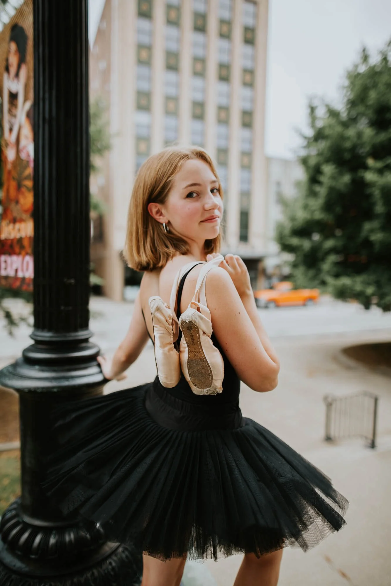 A teen wearing a black tutu and leotard, pointe shoes slung over her should, looks back with a small smile on her lips