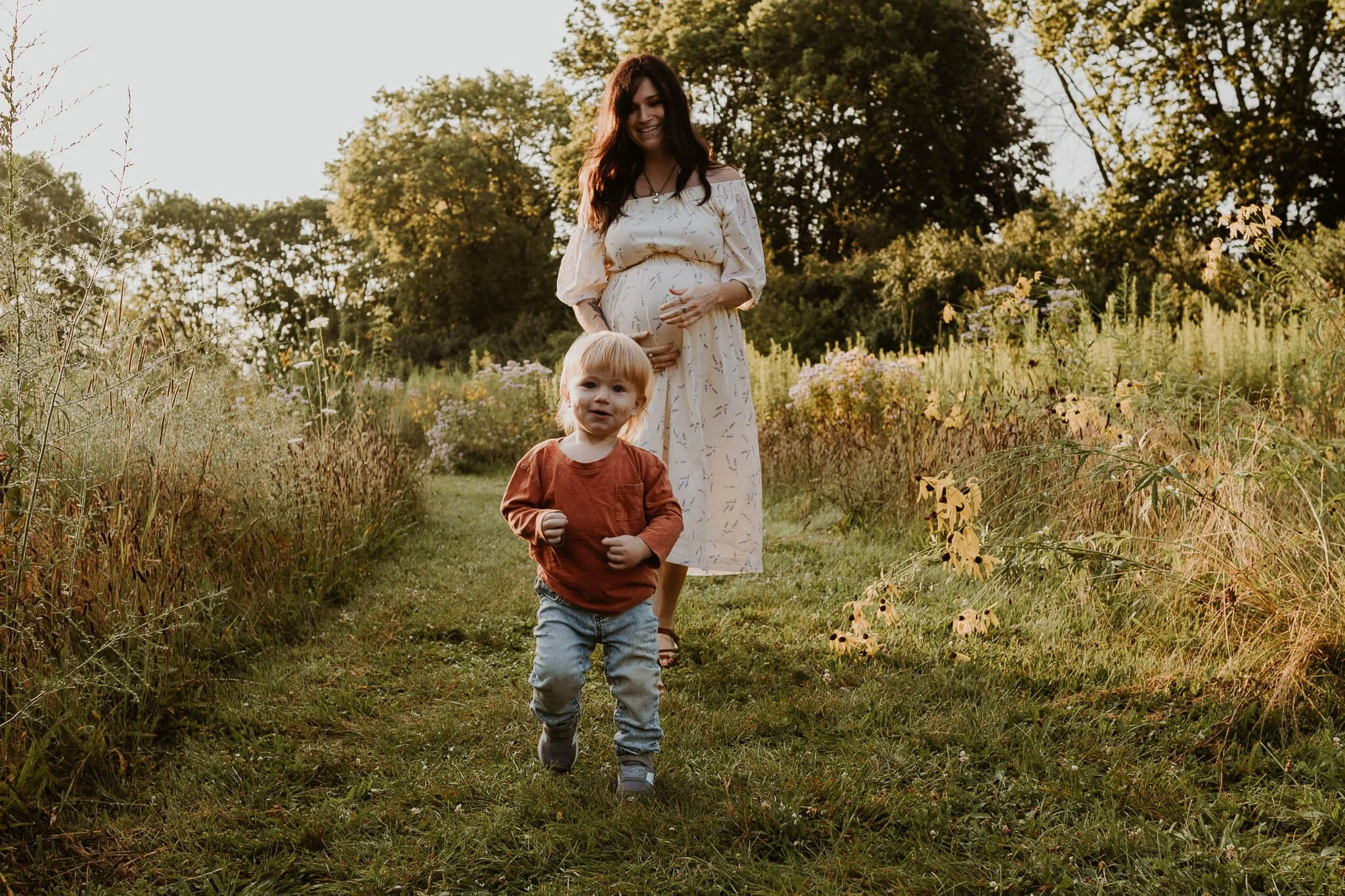 A young boy toddles ahead of his pregnant mom on a mowed path amongst some prairie wildflowers in Bloomington IL