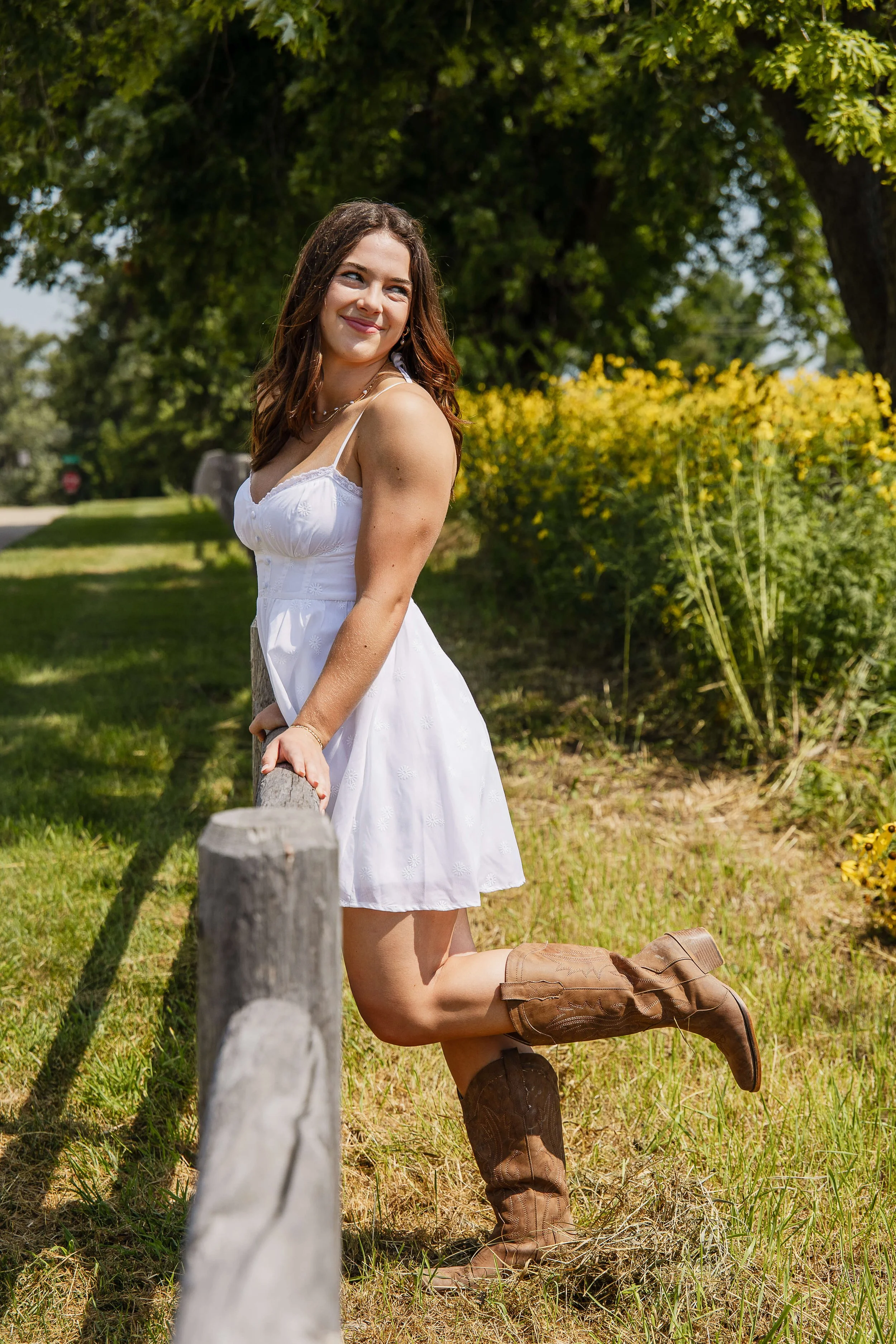A high school senior leans agains a wooden fence post wearing a simple short white dress with her favorite cowboy boots.