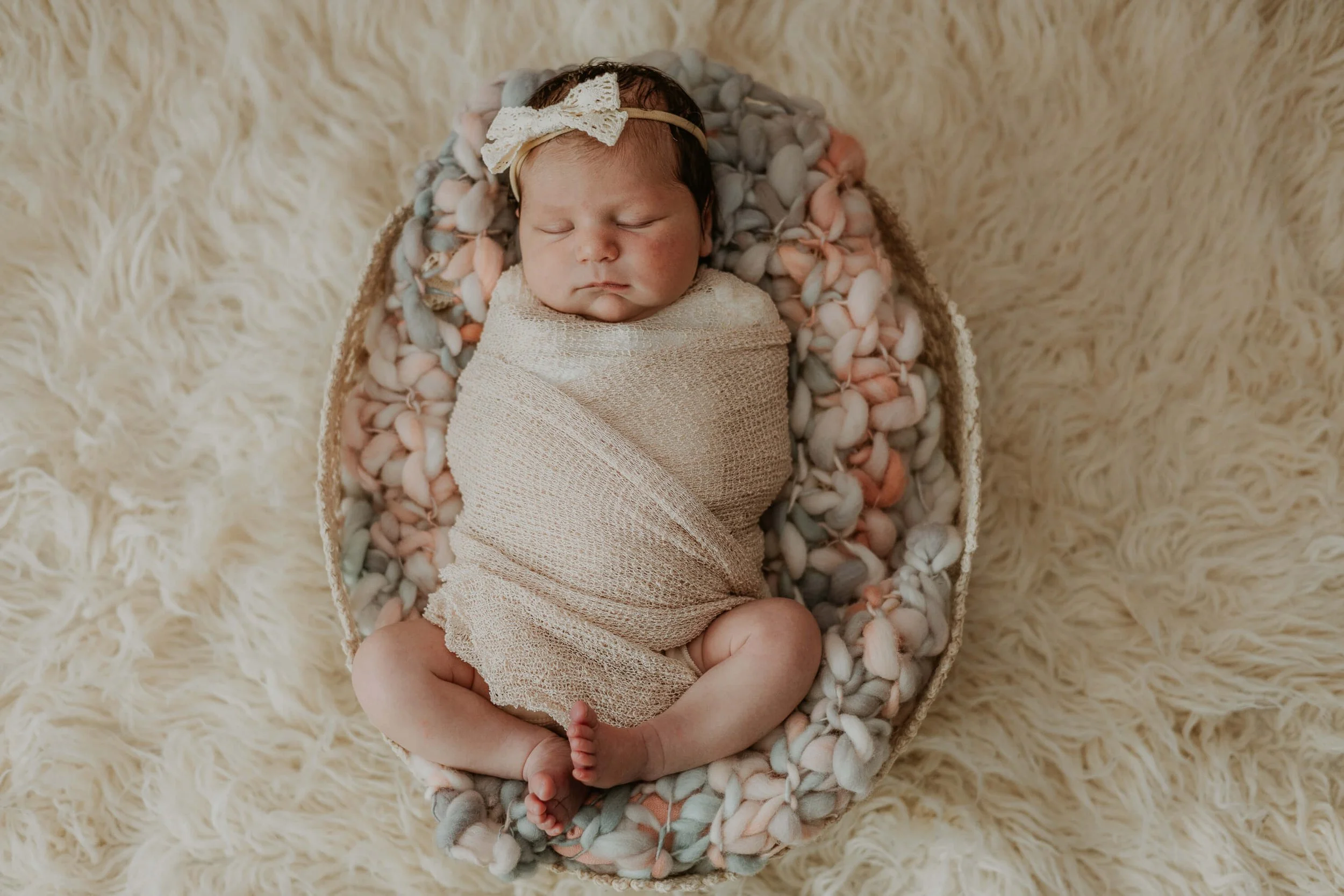 A dark haired baby girl sleeps peacefully while swaddled in a woven basket atop soft woven textures