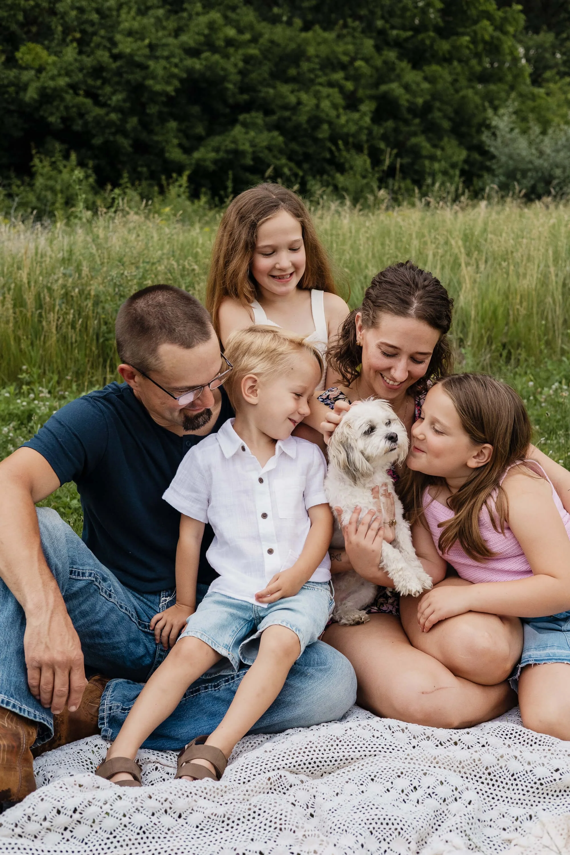 A family of 5 snuggles closely together on a blanket in their yard as they pet and kiss their dog