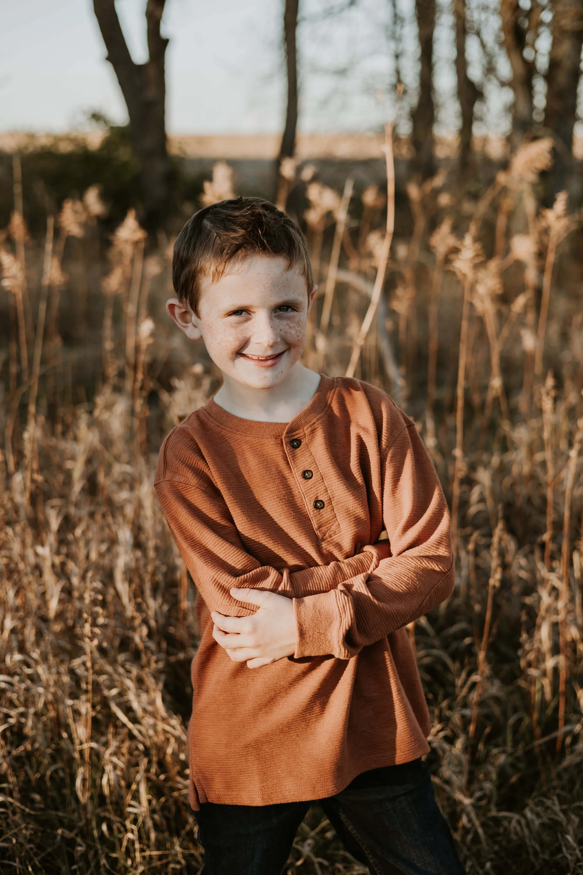 A nine-year-old boy smiles shyly while crossing his arms in front of a line of trees
