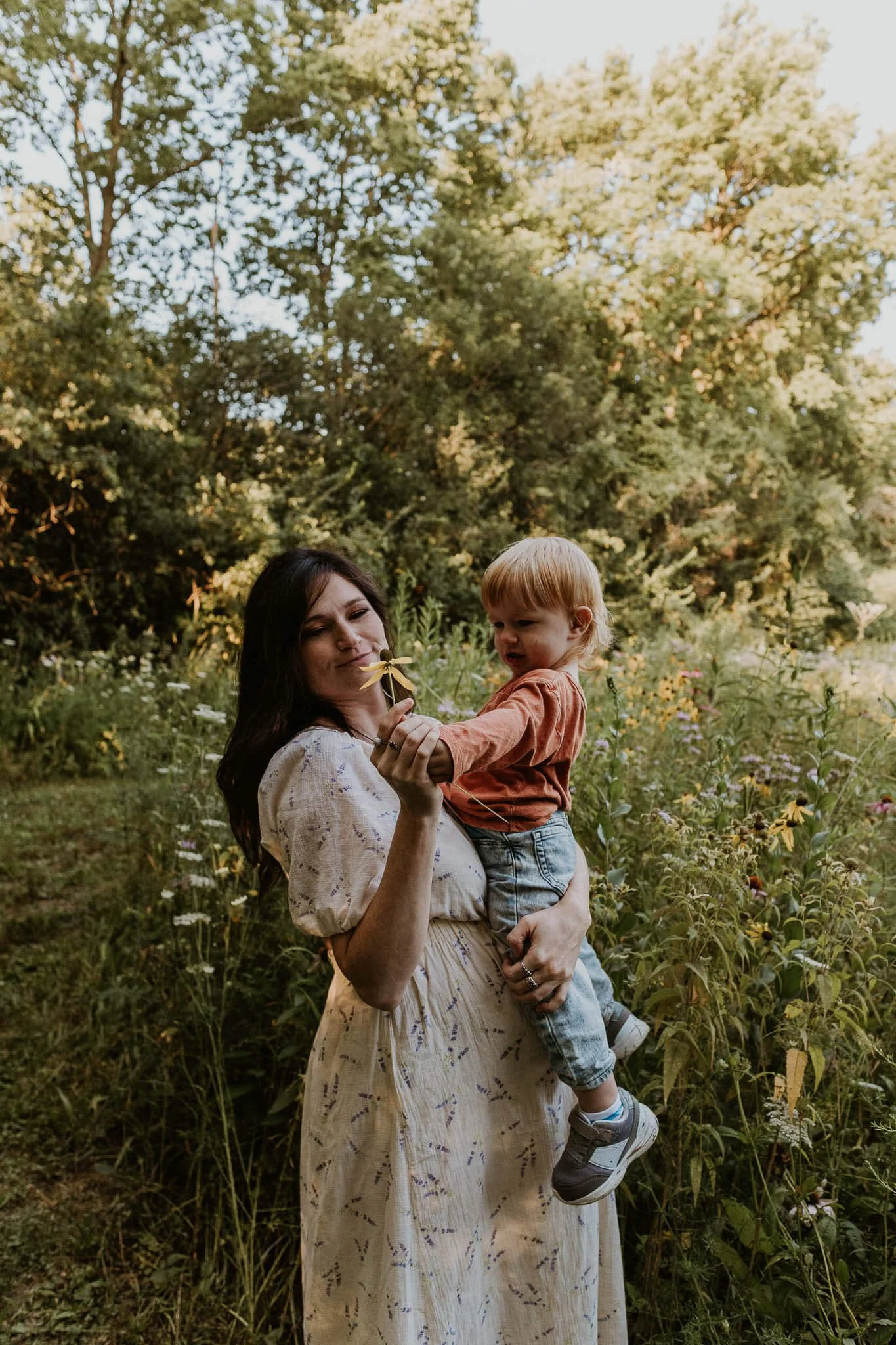 A mother balances her toddler on her growing belly as they discover a flower he picked