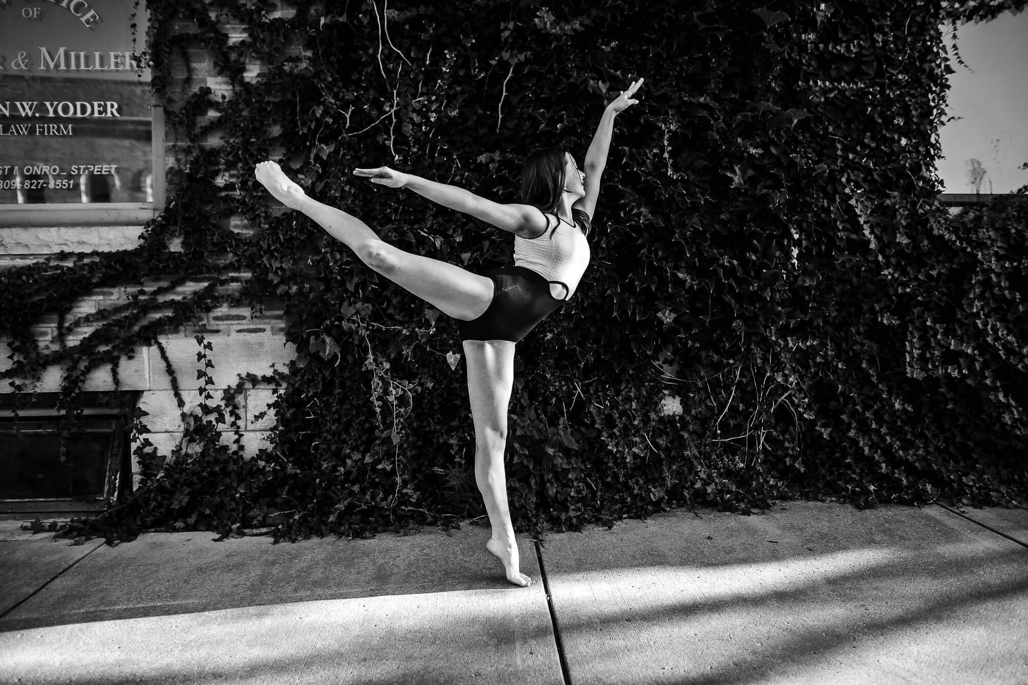 A black and white image of a dancer gracefullly posing outside a building with a wall covered in ivy.