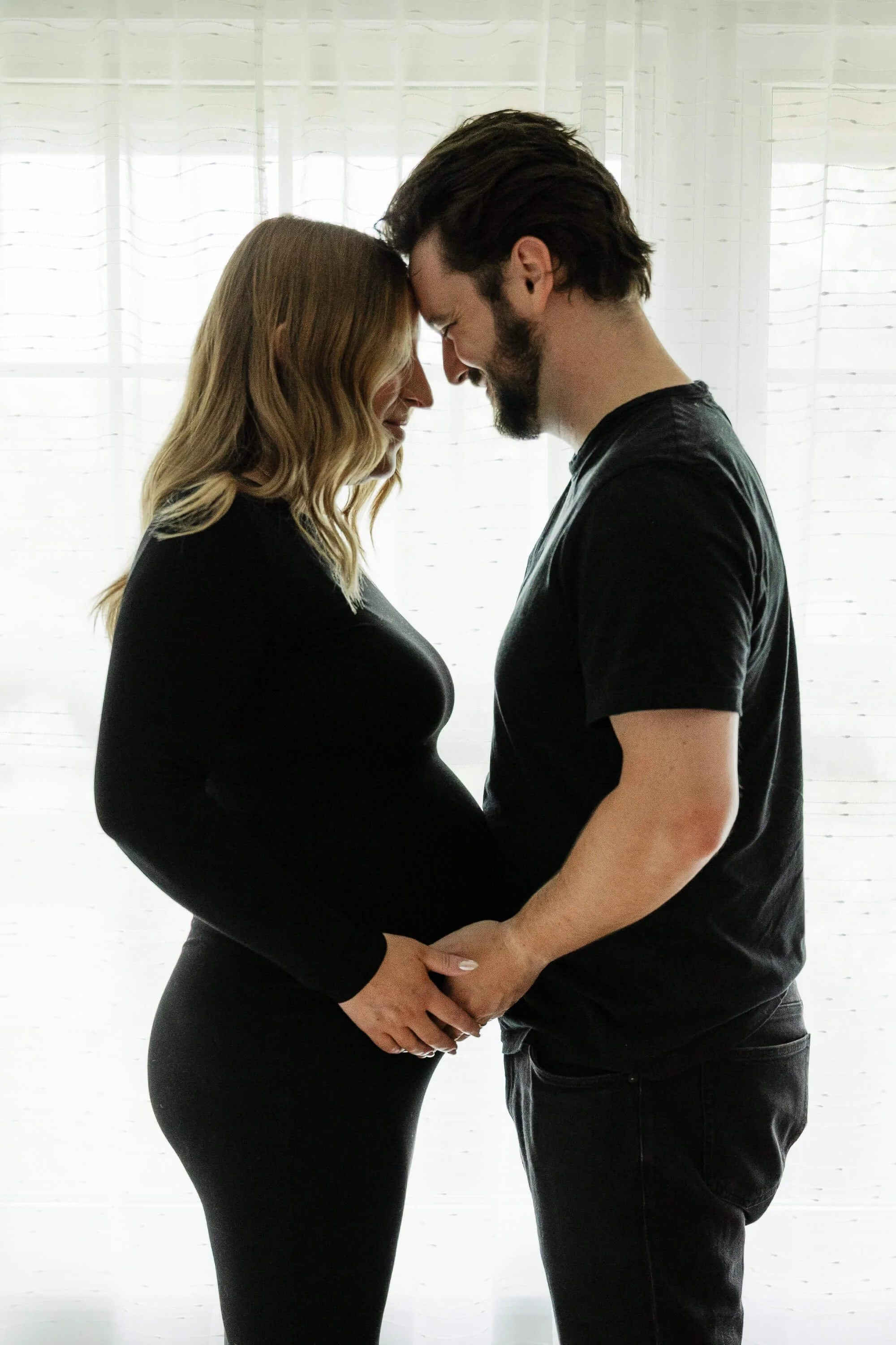 A couple stands forehead to forehead, smiling softly while they both hold the woman's pregnant belly